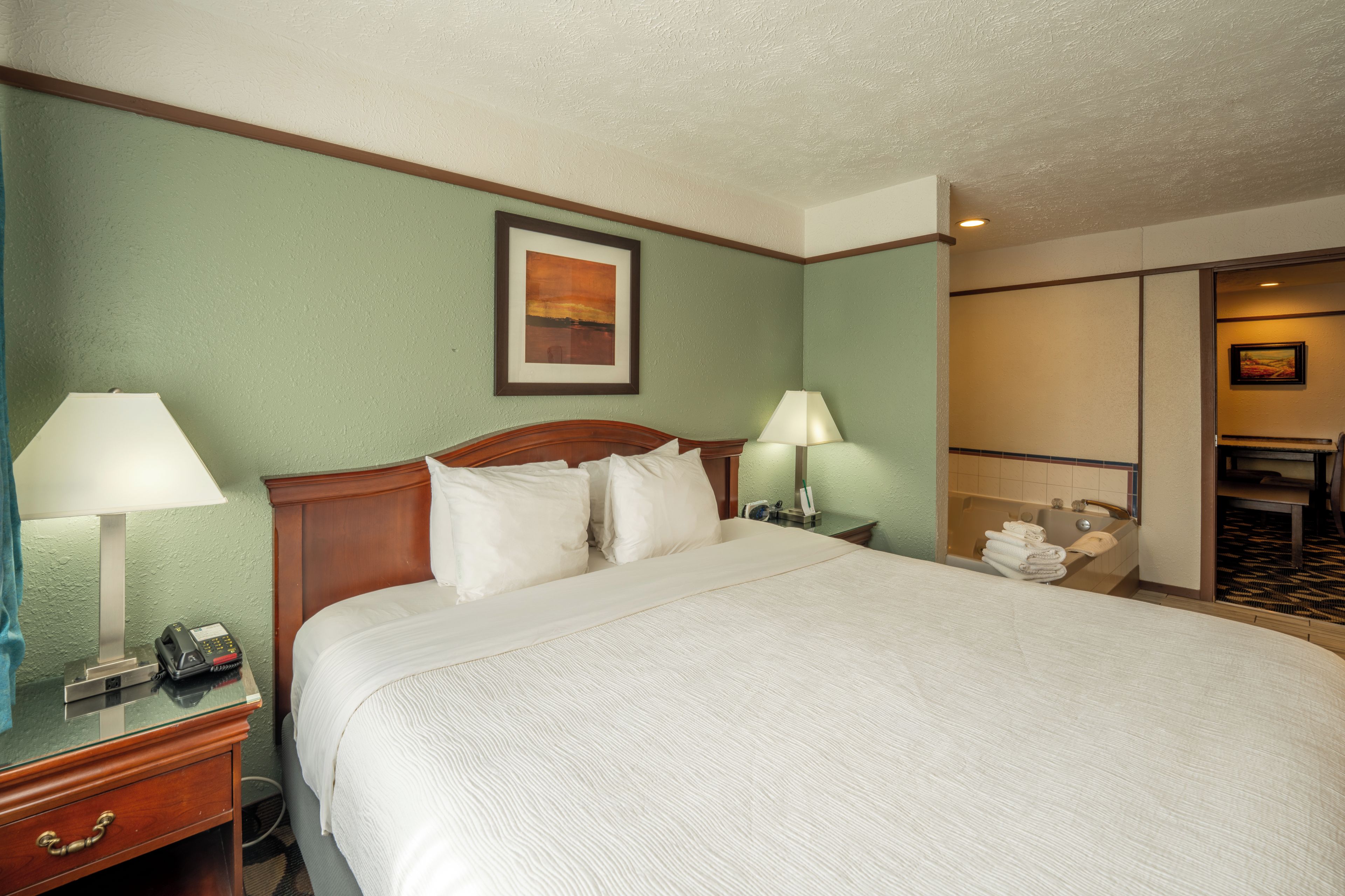 A hotel room with a large bed, two bedside tables with lamps, a painting on the wall, and a bathroom visible through an open door.