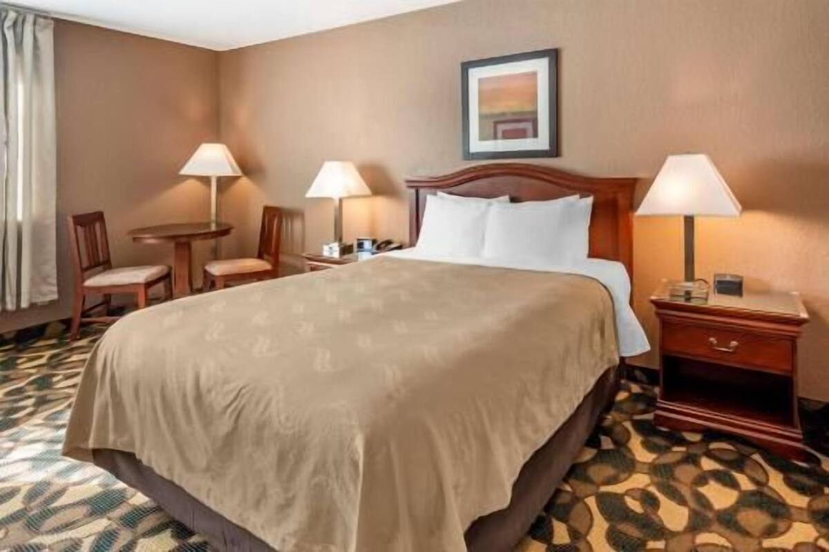 Una habitación de hotel con una cama grande, dos mesitas de noche con lámparas, una silla, una pequeña mesa y un cuadro enmarcado en la pared.