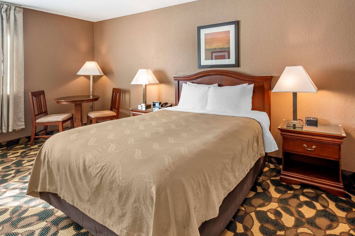Habitación de hotel con una cama grande, dos mesitas de noche, una silla, una mesita pequeña y una lámpara.