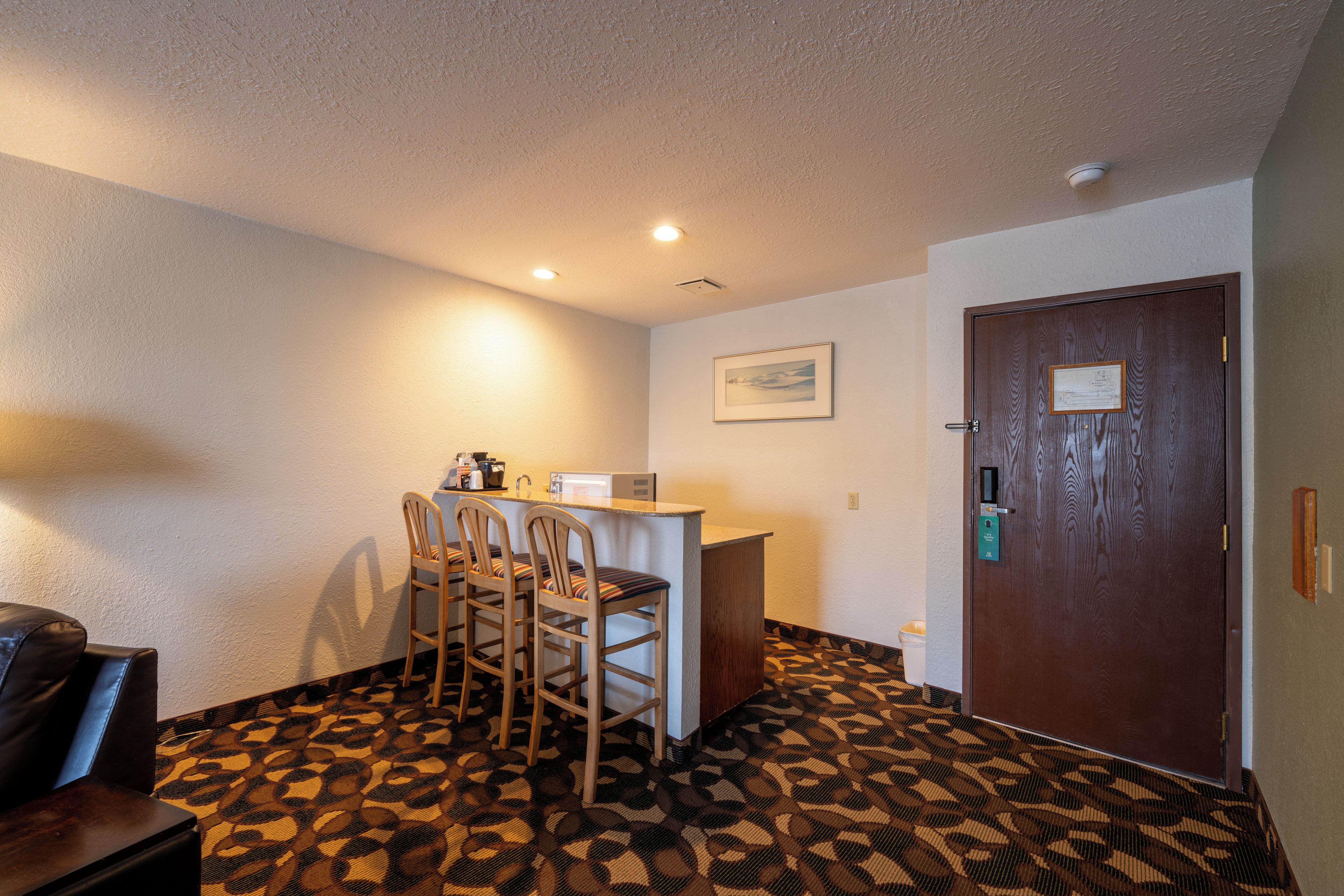 A hotel room with a kitchenette, a dining area with chairs, a patterned carpet, a door with a keycard lock, and a framed picture on the wall.