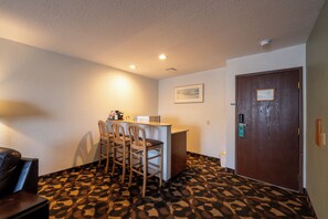 A hotel room with a kitchenette, a dining area with chairs, a patterned carpet, a door with a keycard lock, and a framed picture on the wall. - Quality Inn Paradise Creek Pullman (Pullman)