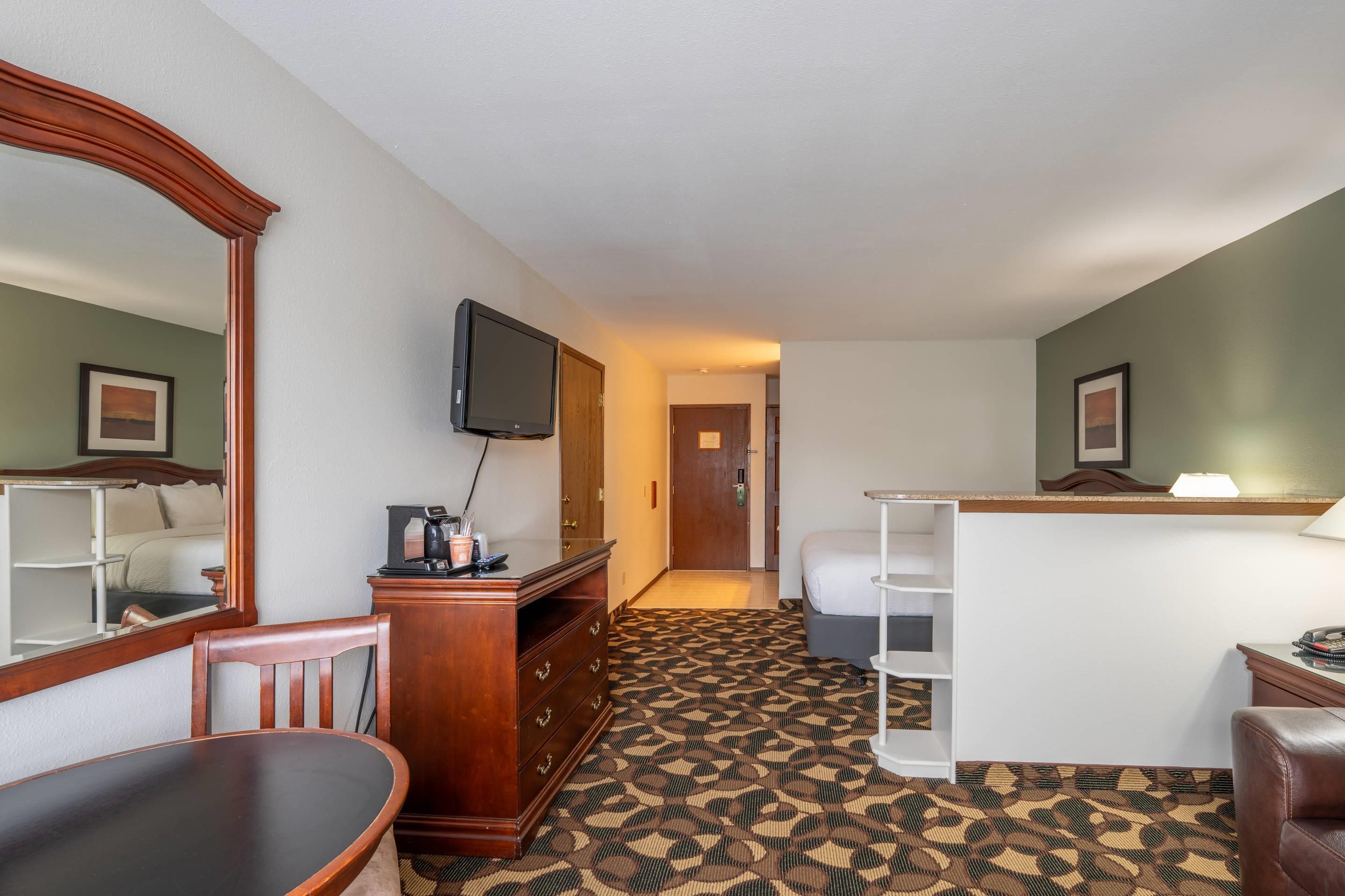 A hotel room with a bed, a desk with a computer, a chair, a dresser, a television, and a patterned carpet.
