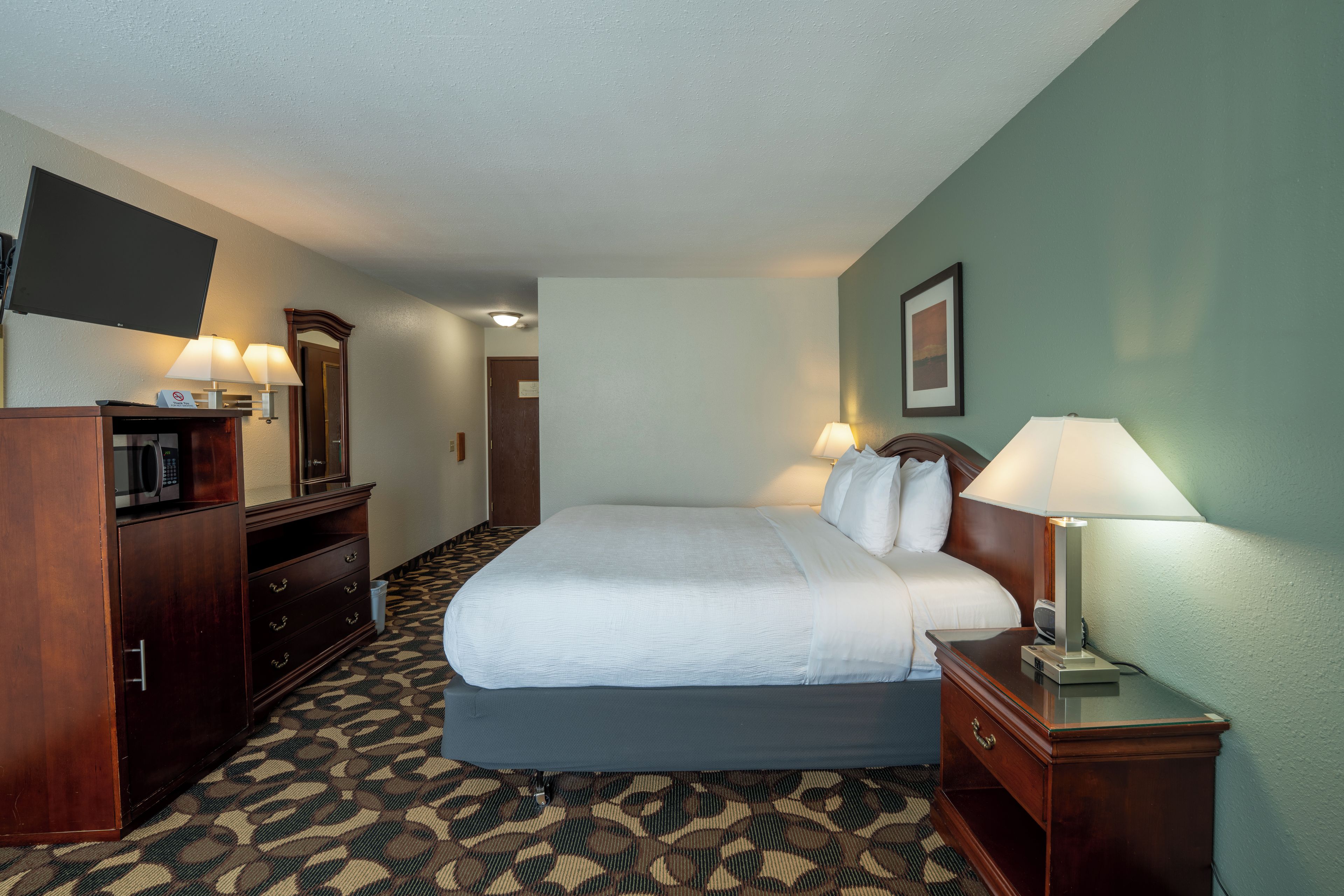 A hotel room with a bed, bedside lamps, a desk, a mirror, a television, and a patterned carpet.