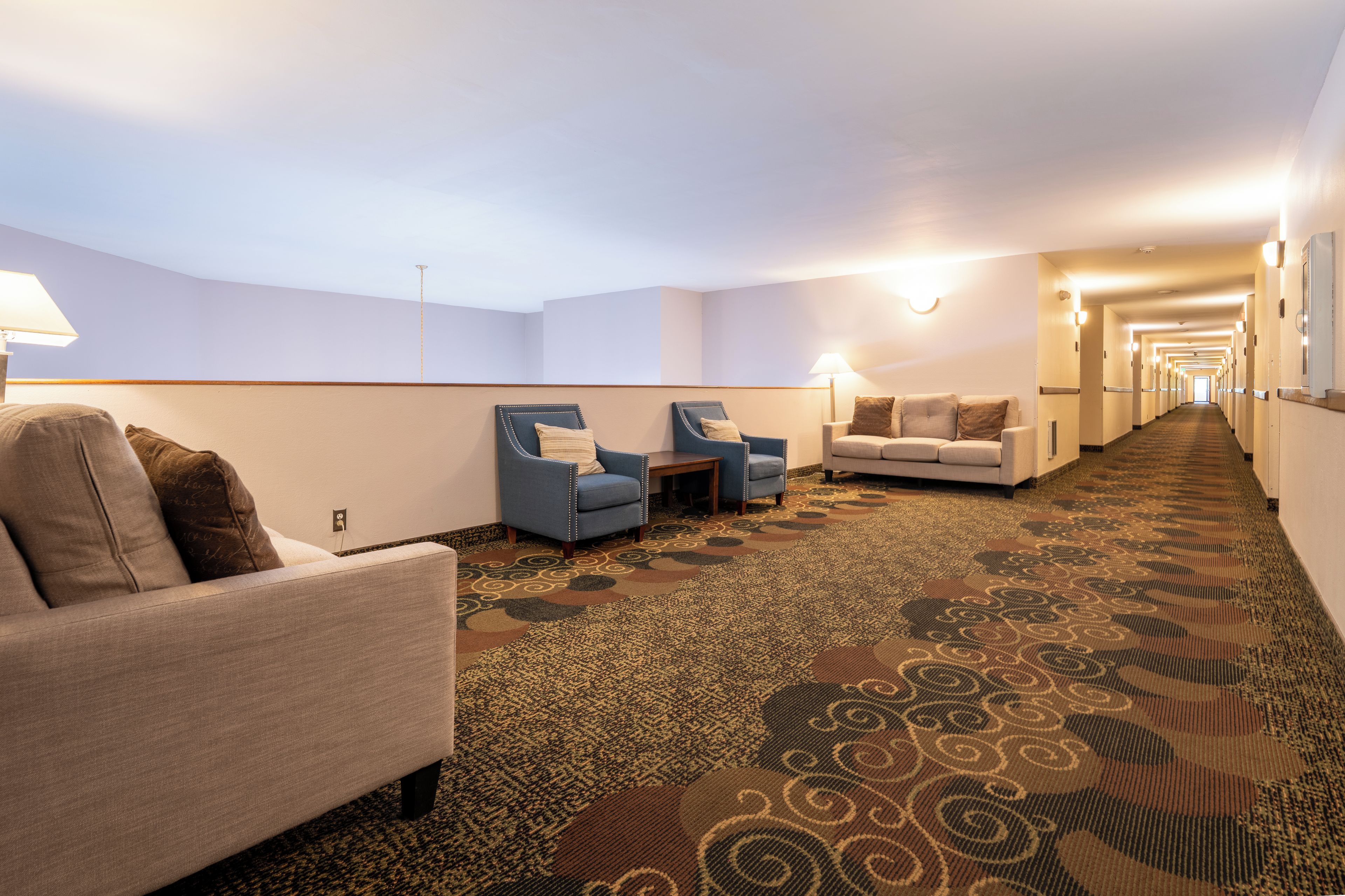 A hotel lobby with a carpeted floor, seating areas with armchairs and a sofa, and a long corridor leading to the exit.