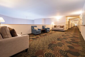 Un vestíbulo de hotel con piso alfombrado, zonas de estar con sillones y un sofá, y un largo pasillo que conduce a la salida.