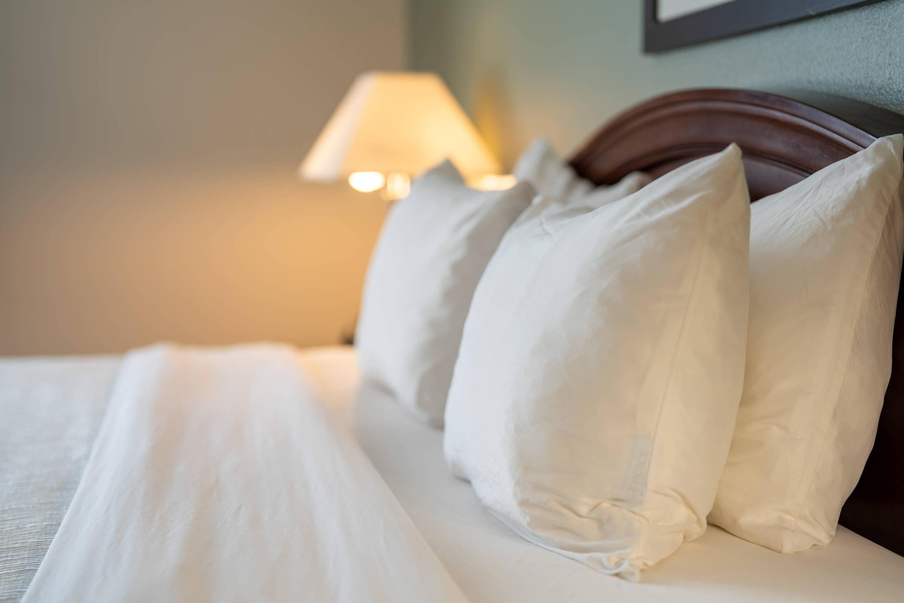 Una cama bien hecha con sábanas y almohadas blancas, un cabecero de madera y una lámpara de pared al fondo.