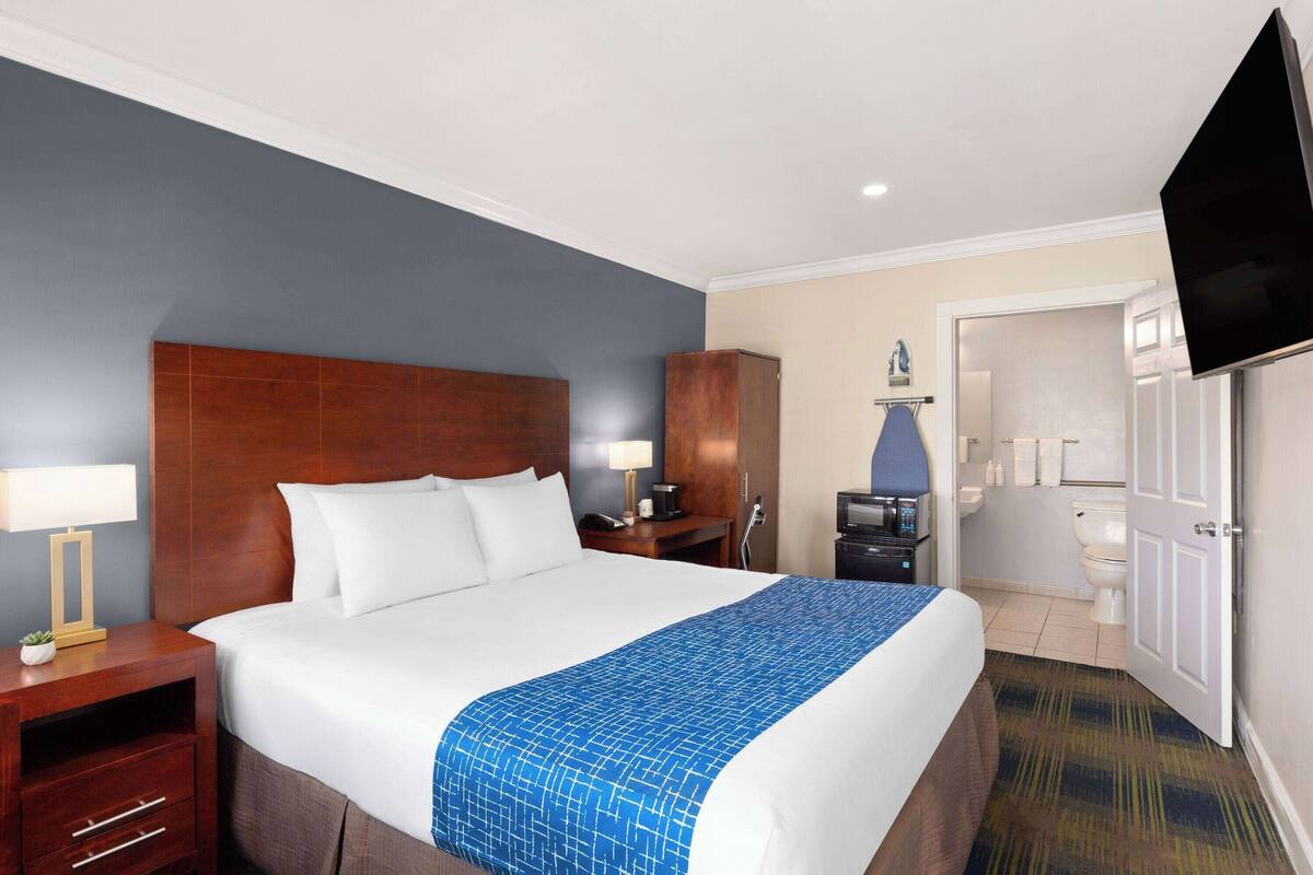 A hotel room with a large bed, a wooden headboard, a TV, a desk with a lamp, a wardrobe, and a bathroom visible through an open door.