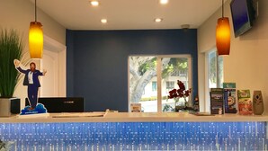 A reception area with a blue patterned counter, a large potted plant, a framed welcome sign, and a mounted television. - Travelodge by Wyndham by Fisherman's Wharf (San Francisco)