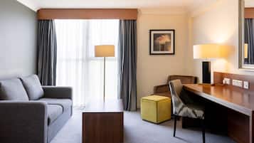 A hotel room with a sofa, a desk, a chair, a small ottoman, and a bed. There is a window with curtains, a lamp, and a framed picture on the wall.