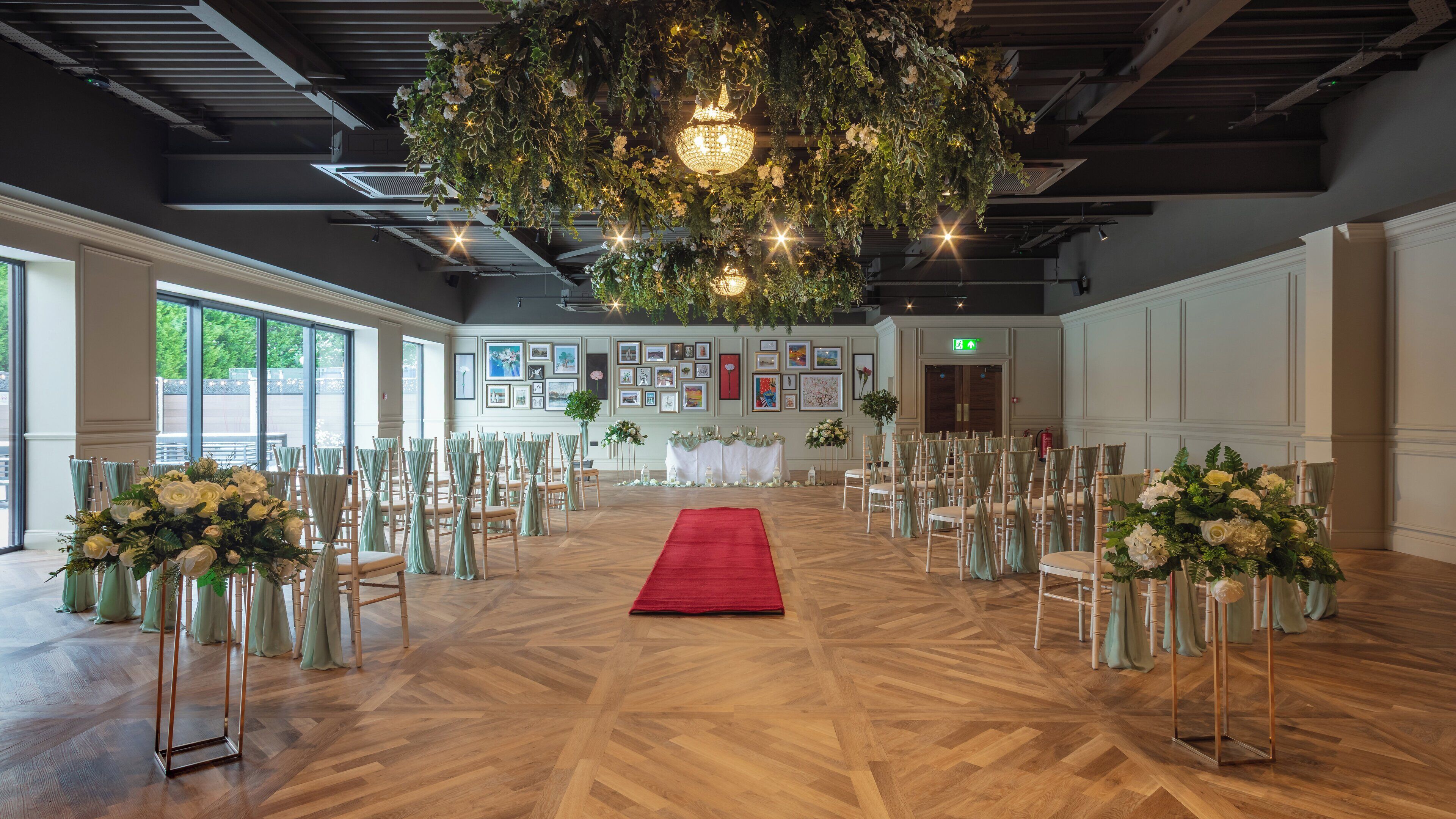 A spacious hall with a red carpet leading to a stage, flanked by rows of chairs with green sashes, and a ceiling adorned with greenery and a chandelier.