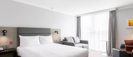 A hotel room with a large bed, a desk with a telephone, a chair, a sofa, and a window with curtains.