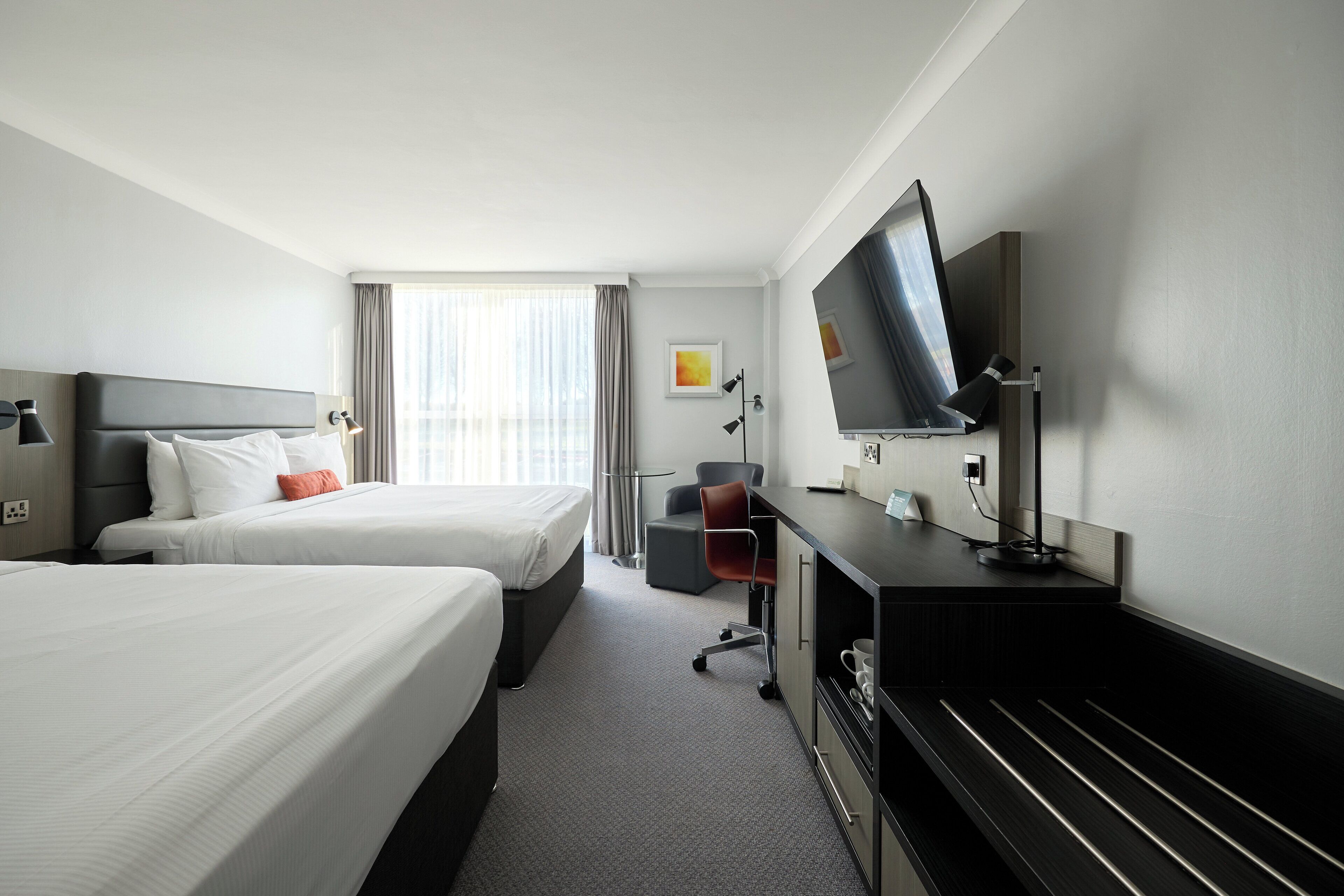A hotel room with two beds, a desk with a flat-screen TV, a chair, and a large window with curtains.