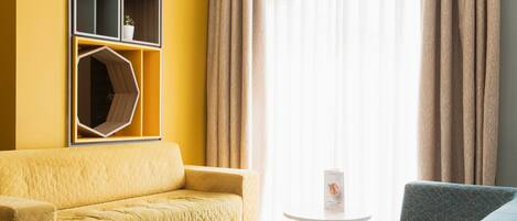 A room with a yellow sofa, a blue armchair, a small round table, and a wall-mounted shelf with a potted plant.