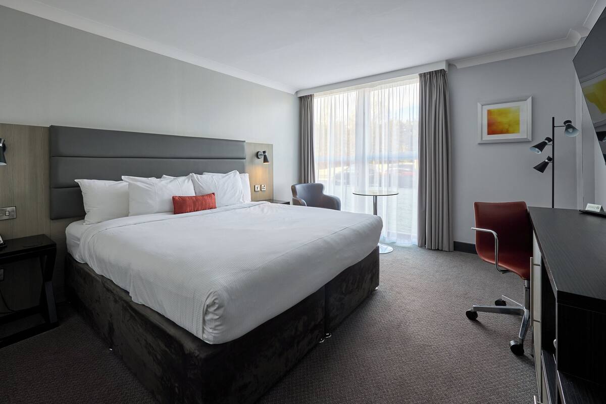 A hotel room with a large bed, a desk, a chair, and a view of the city through the window.