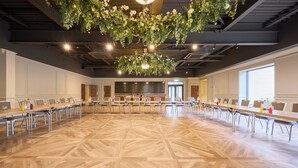Sala de conferência com amplo piso de madeira polida e longas mesas dispostas para reuniões.