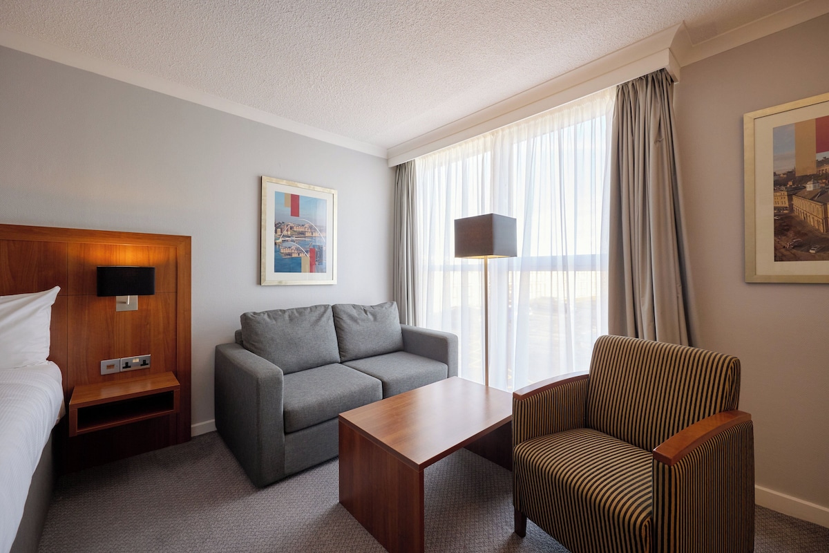 A hotel room with a bed, a grey sofa, a striped armchair, a wooden nightstand, a table, and a large window with curtains.