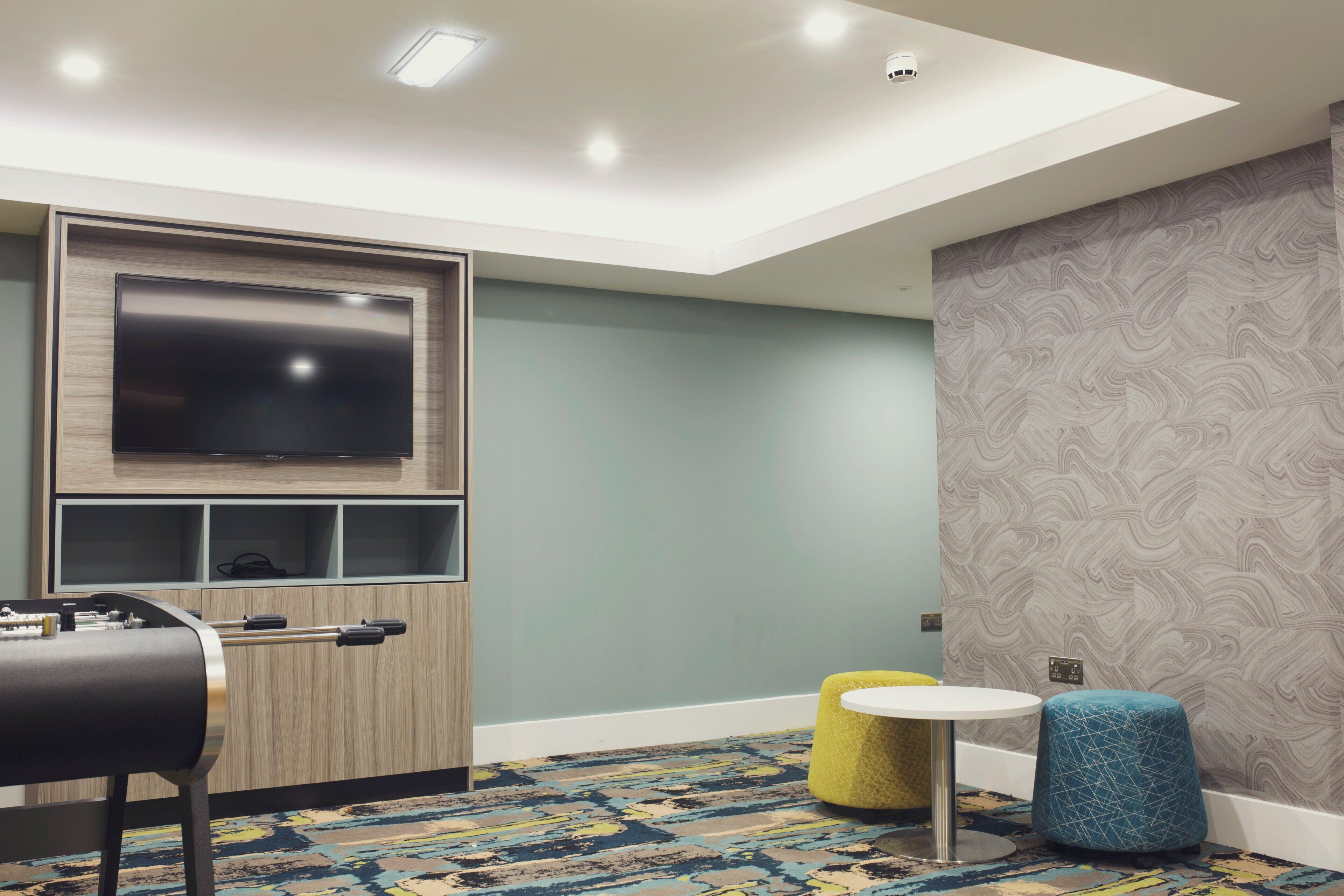 A modern room with a flat-screen TV mounted on a wooden cabinet, a small round table, two colorful ottomans, and a patterned carpet.