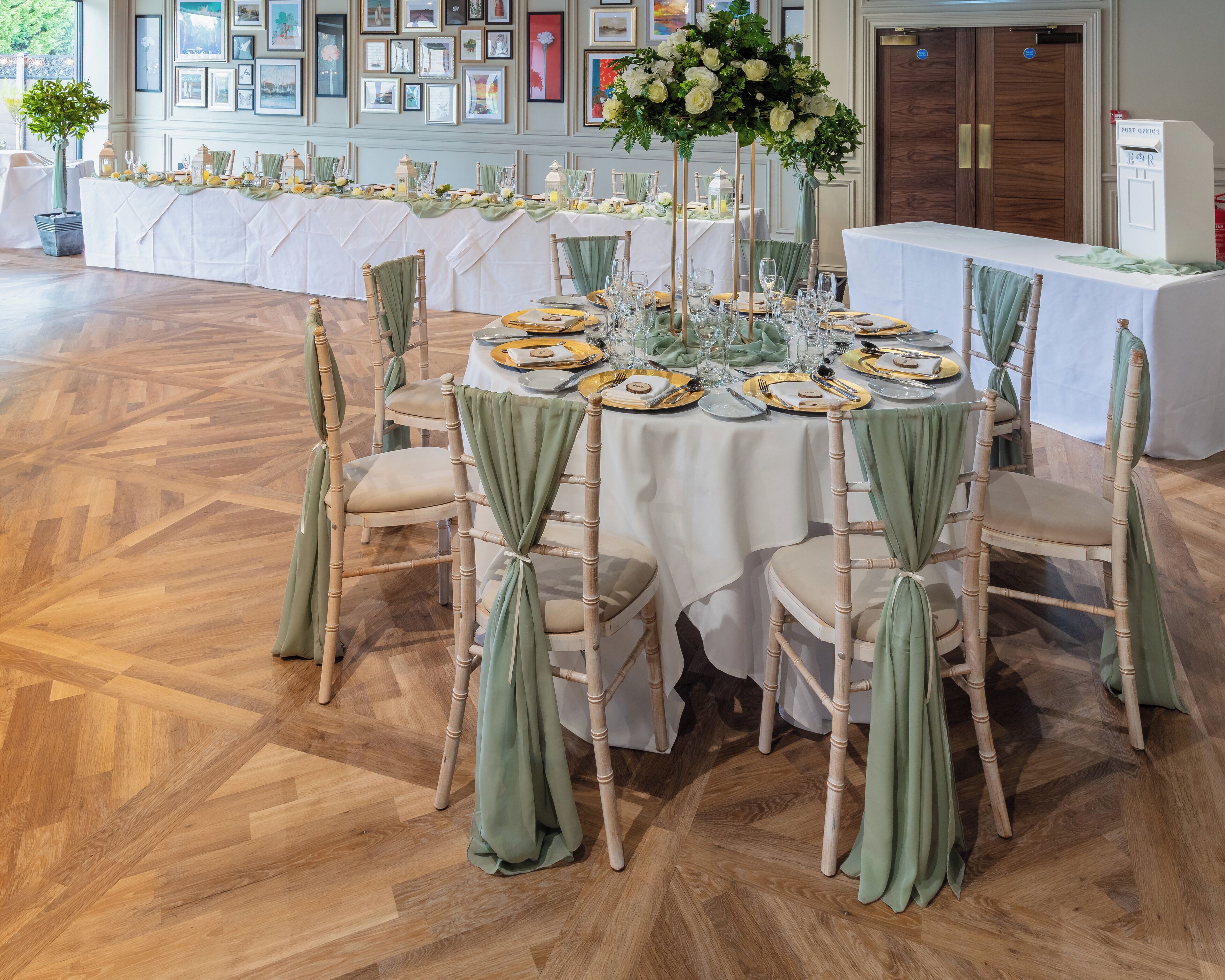 Una mesa redonda dispuesta para una comida, con sillas cubiertas de tela verde, un centro de mesa floral y una pared decorada con cuadros.