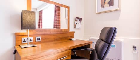A wooden desk with a mirror, a black office chair, a lamp, and a framed picture on the wall.