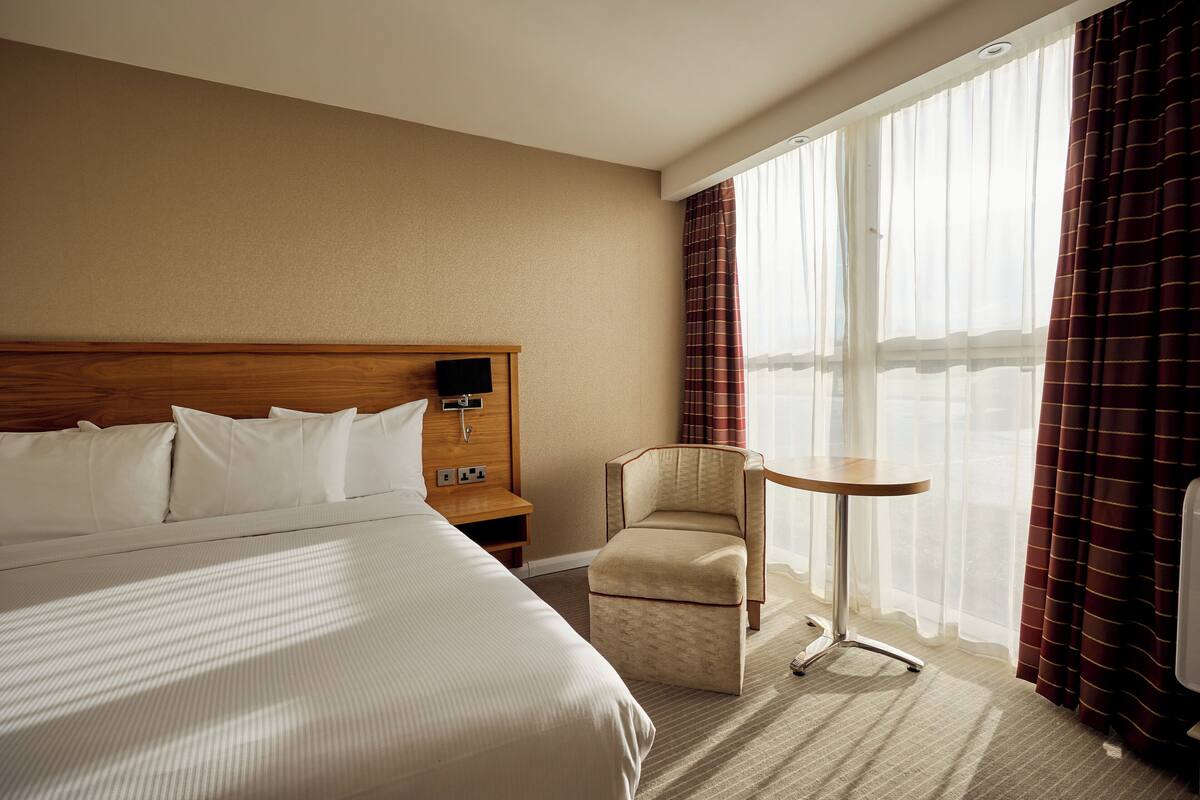 A hotel room with a large bed, a wooden headboard, a small table, a chair, and a view of the water through sheer curtains.