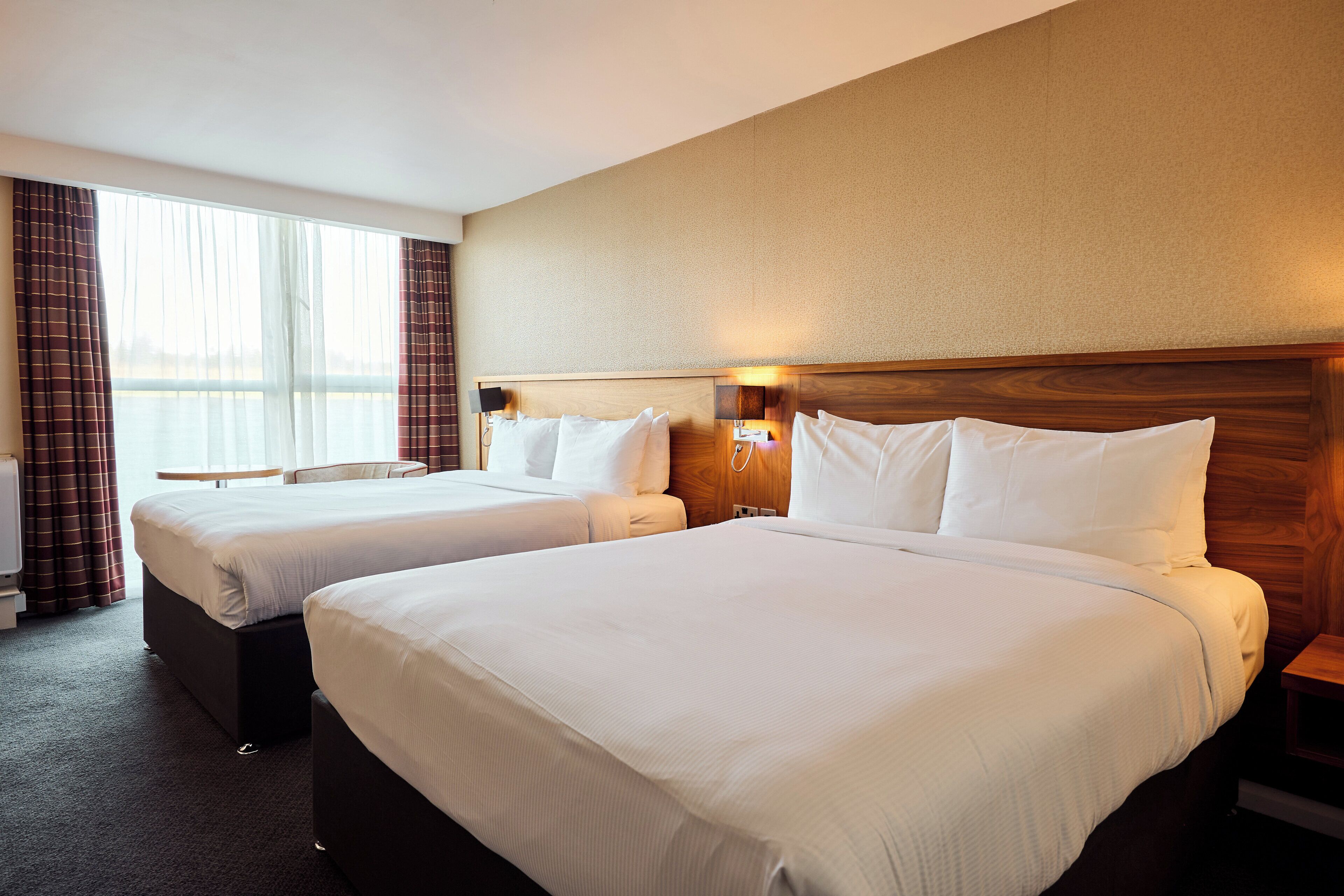 A hotel room with two beds, a wooden headboard, a nightstand, and a window with curtains.