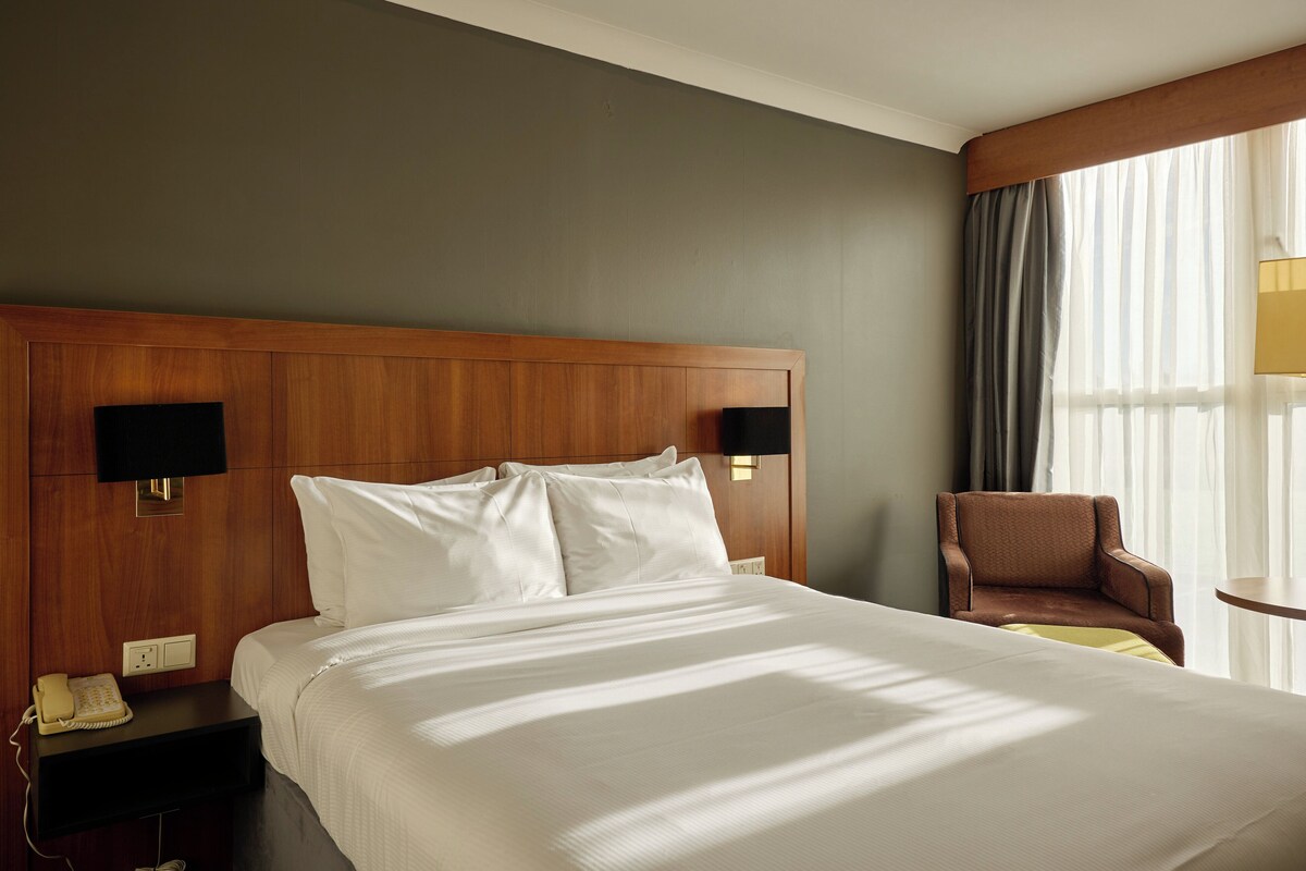 A hotel room with a large bed, a wooden headboard, two bedside tables with lamps, a chair, and a window with curtains.