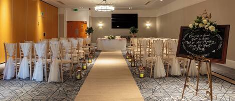 A hotel room with a long beige carpet, wooden chairs with white covers, a chandelier, and a sign with a floral arrangement.