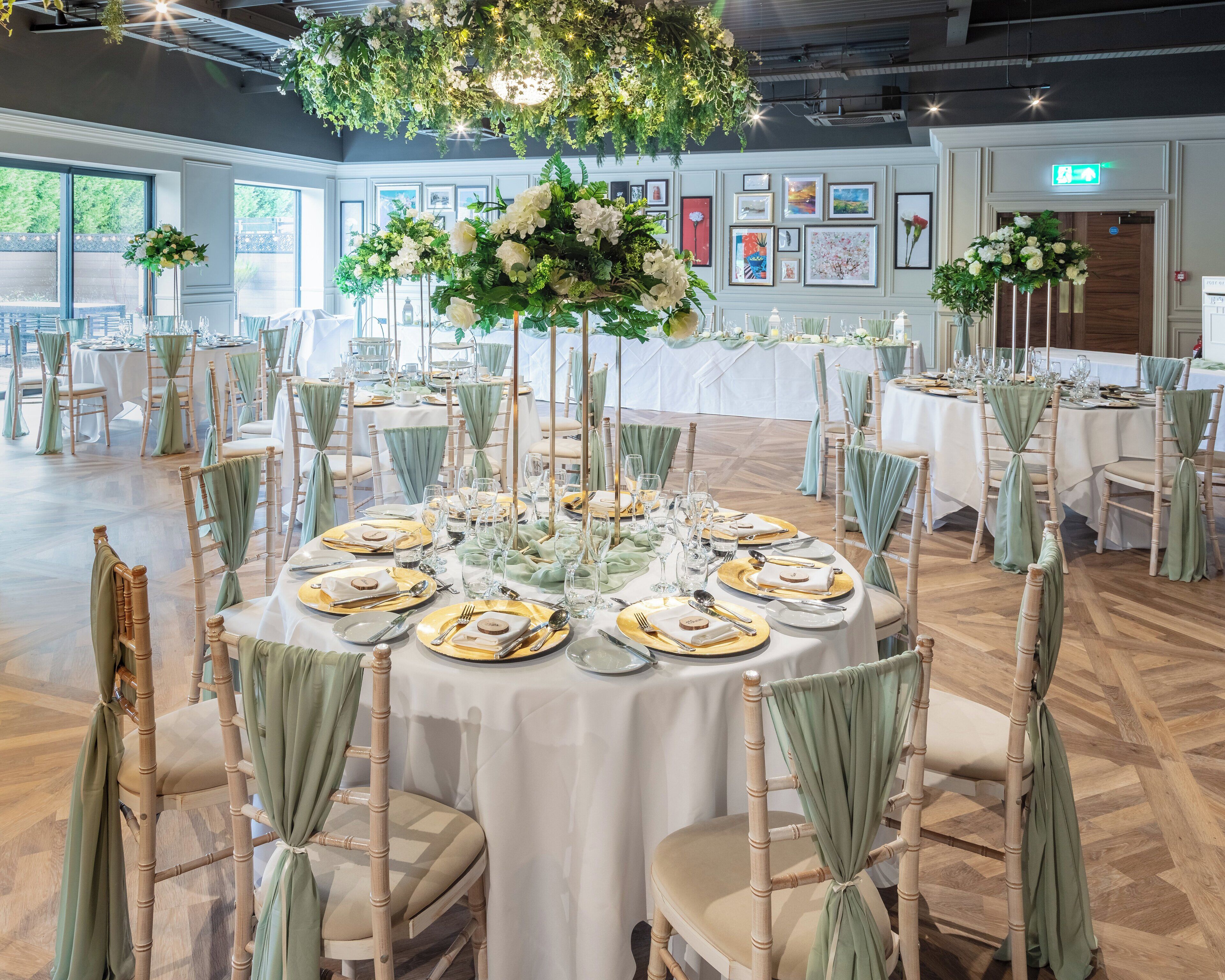 Un salón de banquetes con mesas redondas dispuestas para un evento formal, con manteles blancos, platos subeplato dorados y fundas de silla verdes.