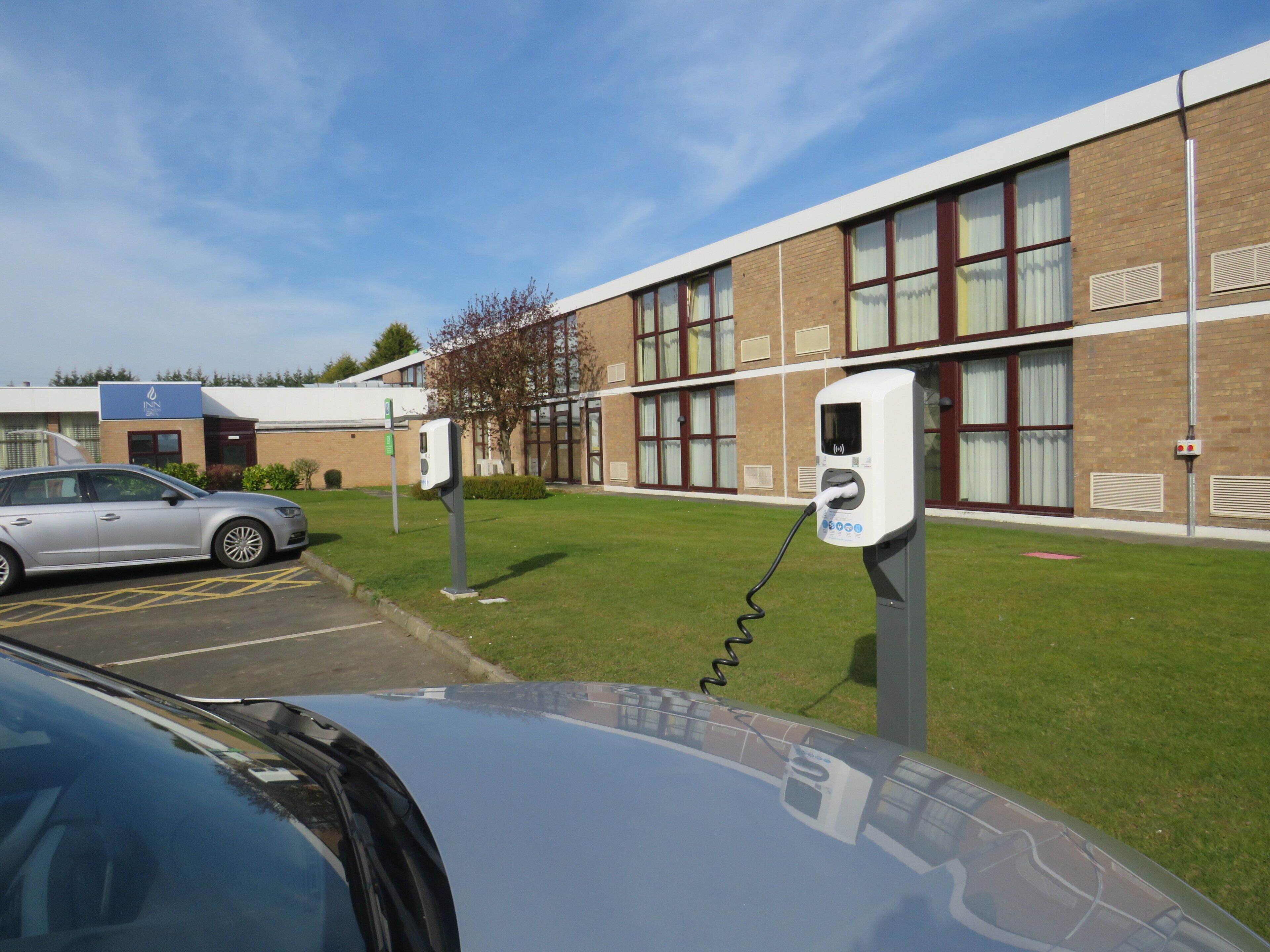 Un espace de stationnement équipé de bornes de recharge pour véhicules électriques, un bâtiment doté de grandes fenêtres et une voiture garée au premier plan.