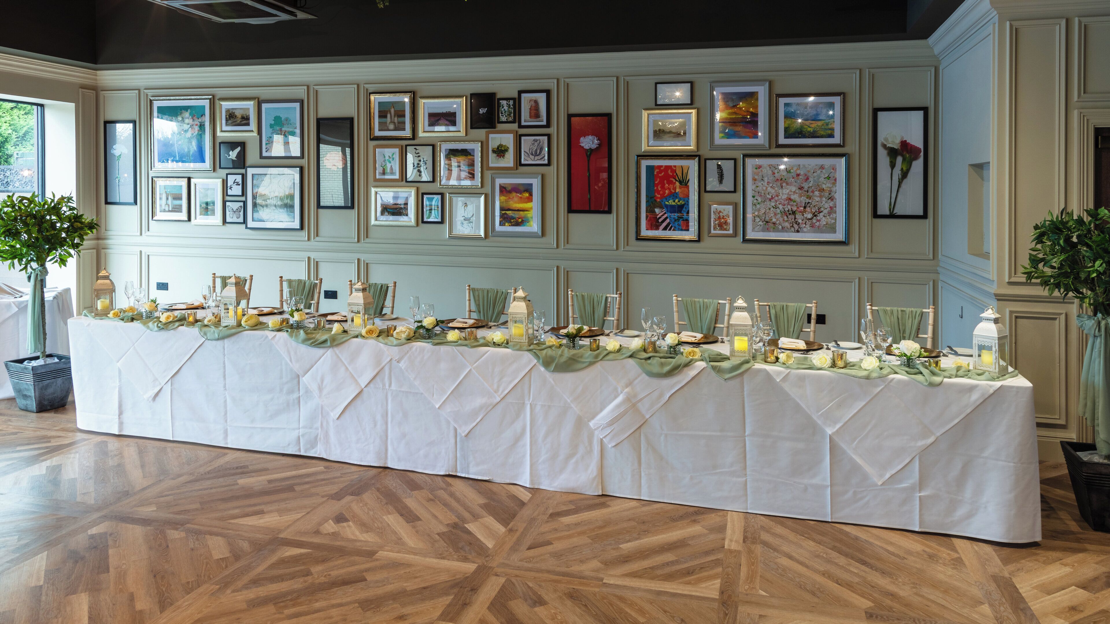A long dining table set with white tablecloths, candles, and floral centerpieces, surrounded by framed artworks on the walls.