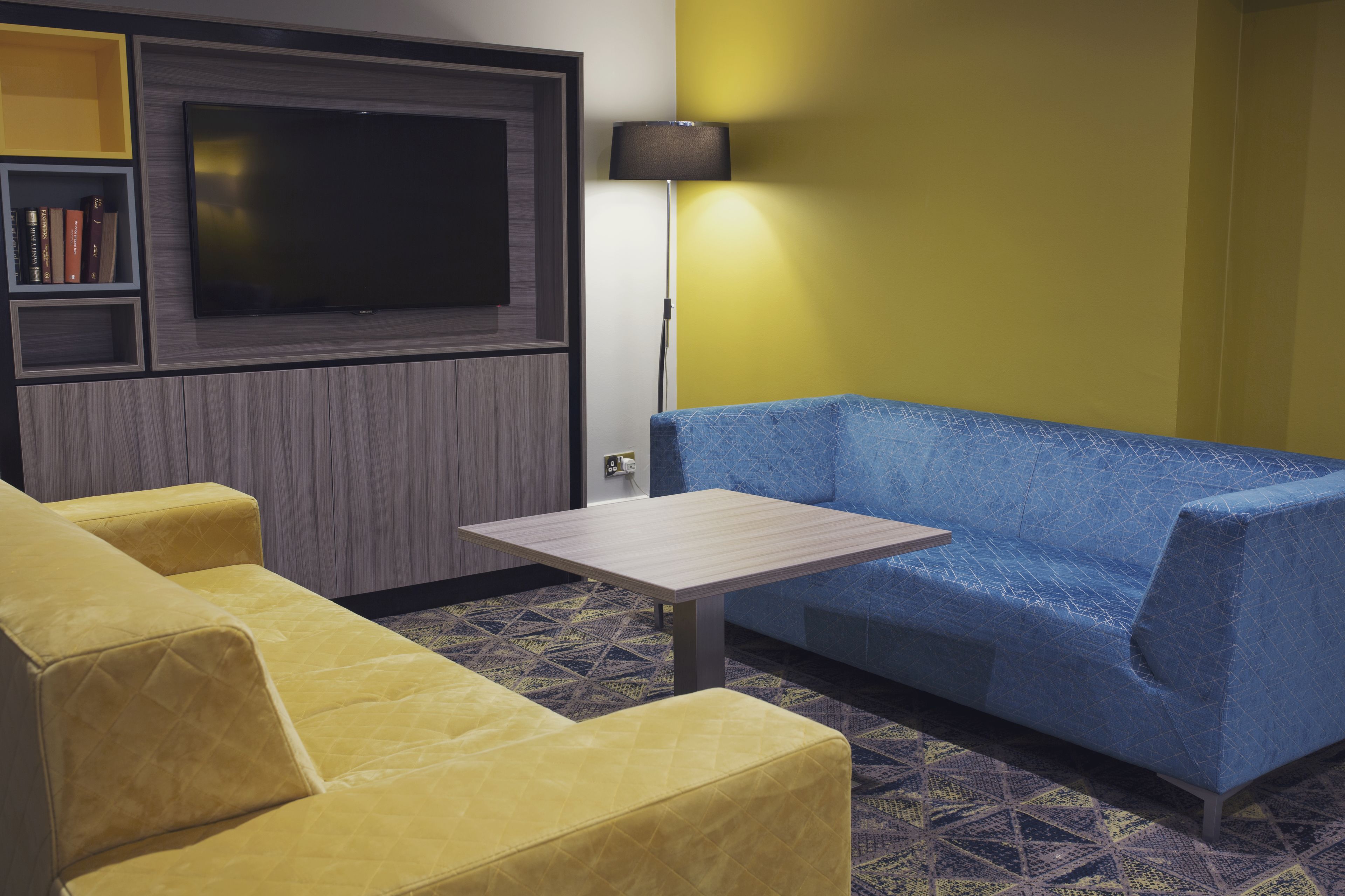Een kamer met een groot flatscreen televisietoestel aan de muur, een blauwe bank, een gele bank en een kleine tafel.