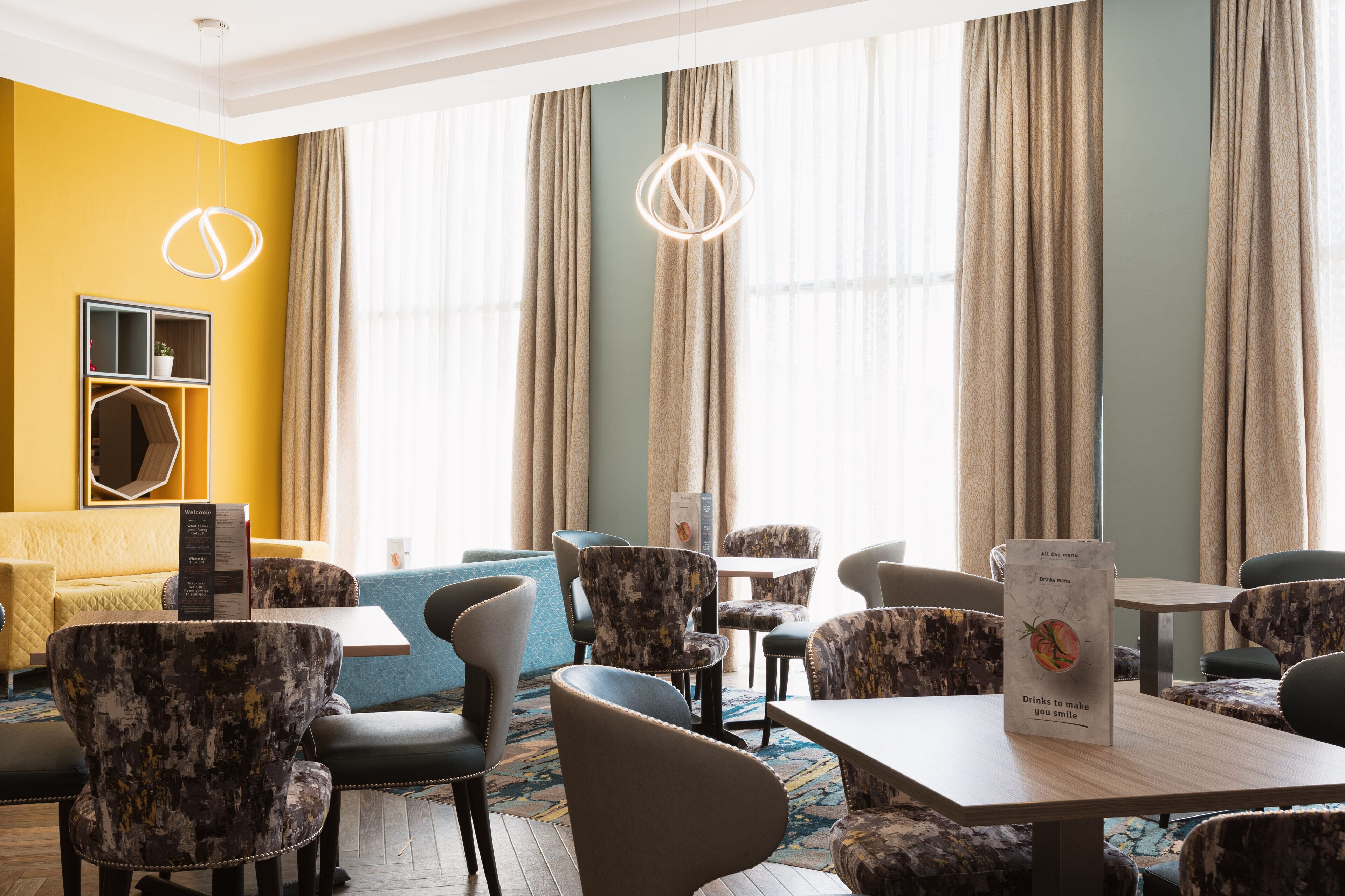 Un hall d’hôtel avec un mur jaune, une grande fenêtre avec des rideaux et différents types de sièges, notamment des canapés et des fauteuils.