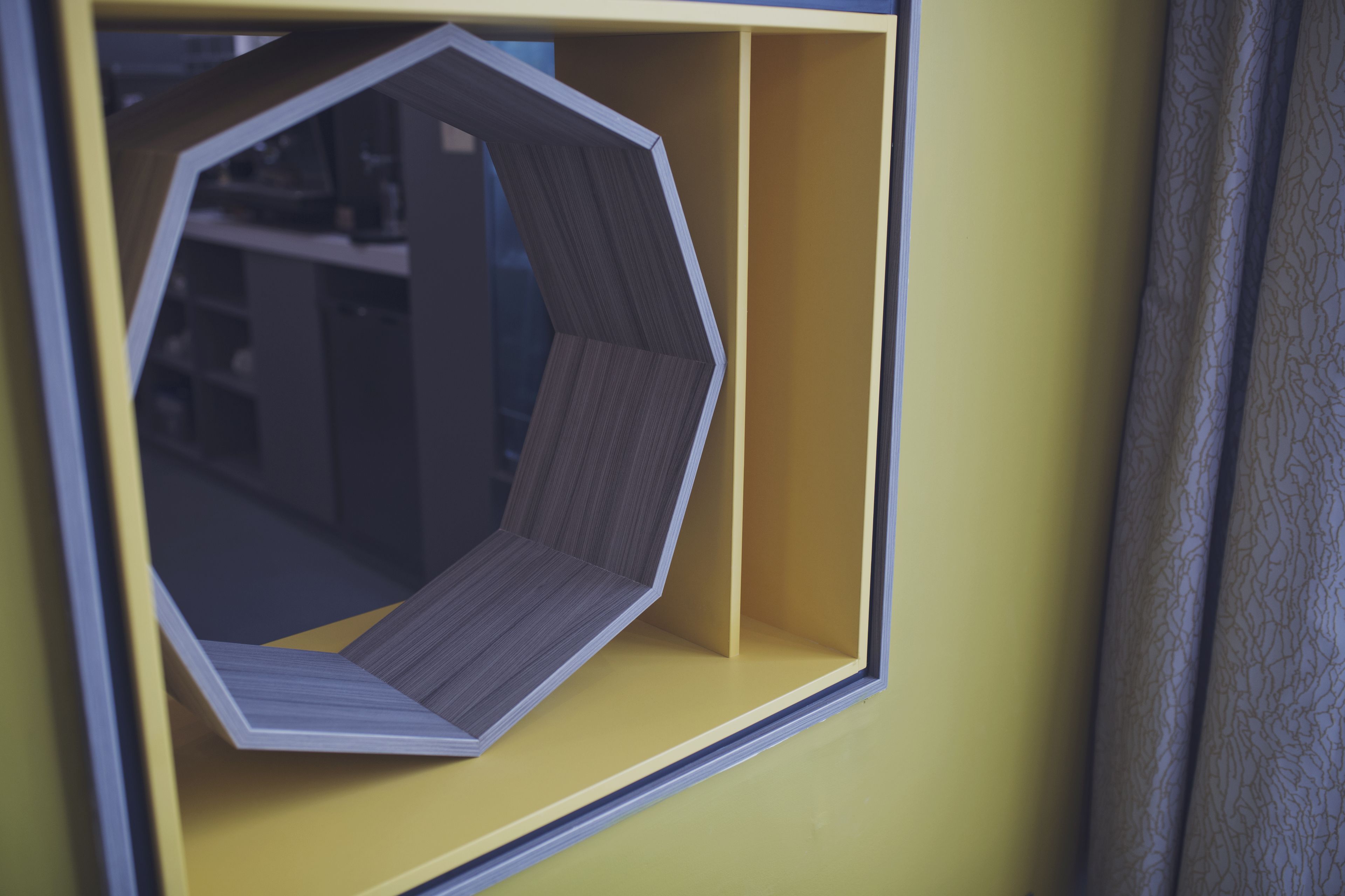 A hexagonal window with a wooden interior, framed in black, against a yellow wall.