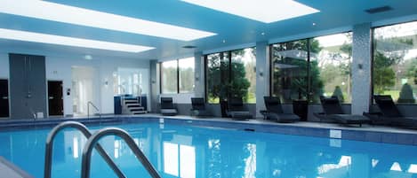 Una piscina interior con agua azul cristalina, rodeada de sillas de descanso y una escalera que conduce a un nivel superior.