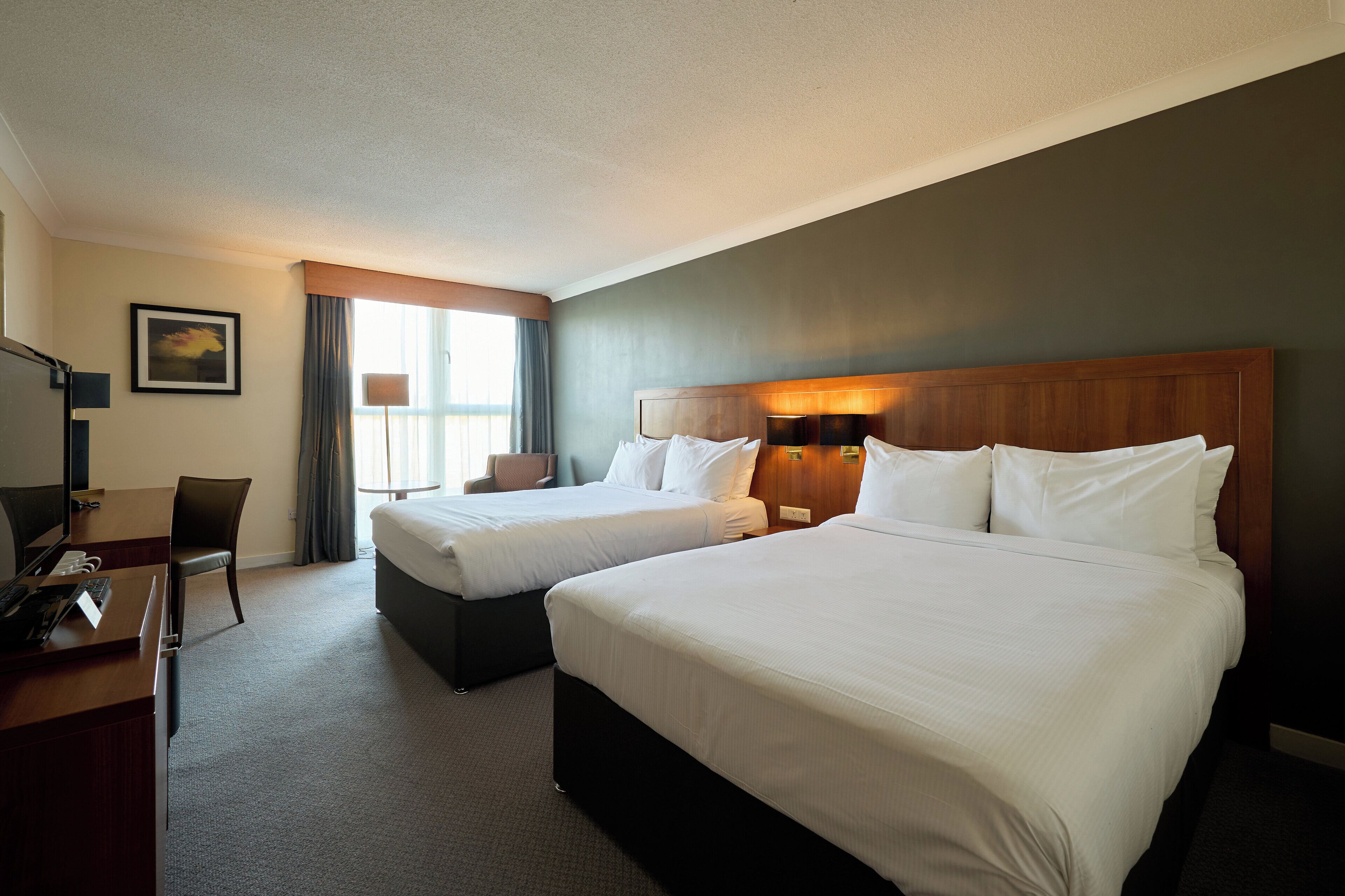 Una habitación de hotel con dos camas, un escritorio con computadora, una silla y una ventana con cortinas.