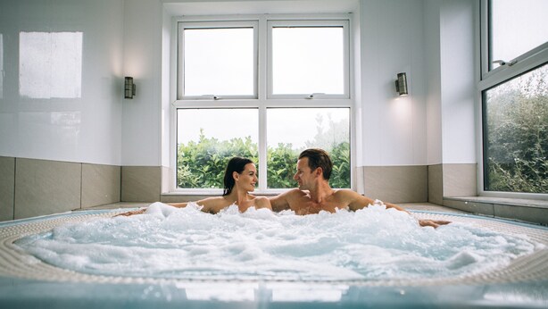 Um casal em uma banheira de hidromassagem, com vista para o verde lá fora.