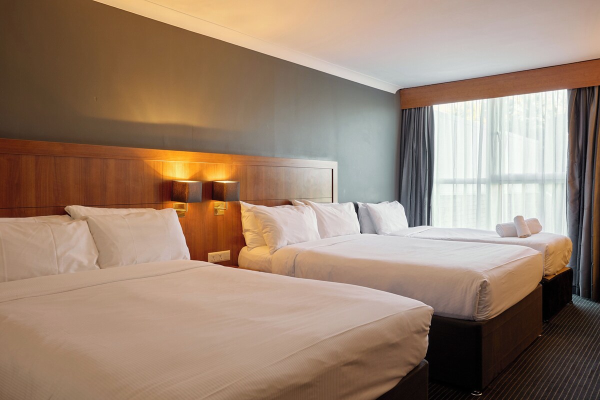 A hotel room with two beds, a wooden headboard, a wall-mounted light, and a large window with curtains.