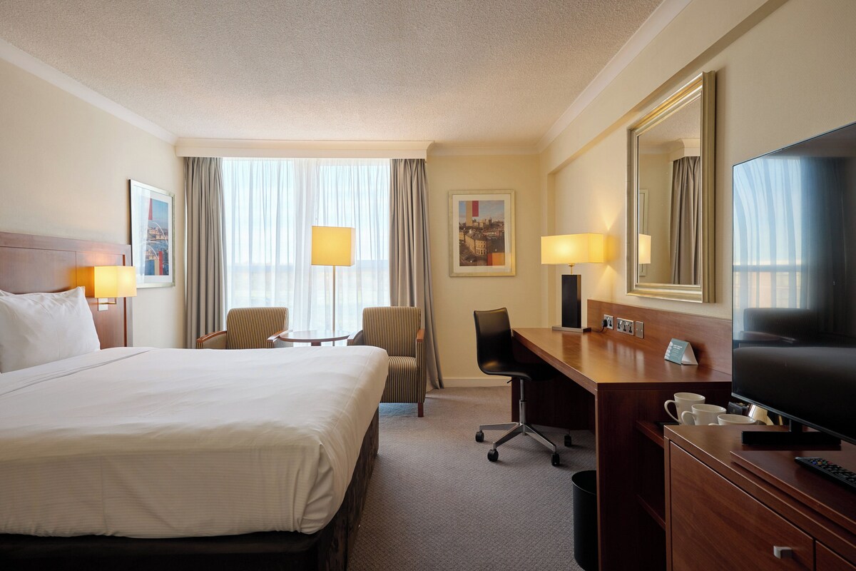 A hotel room with a large bed, a desk with a TV, a chair, and a desk lamp.