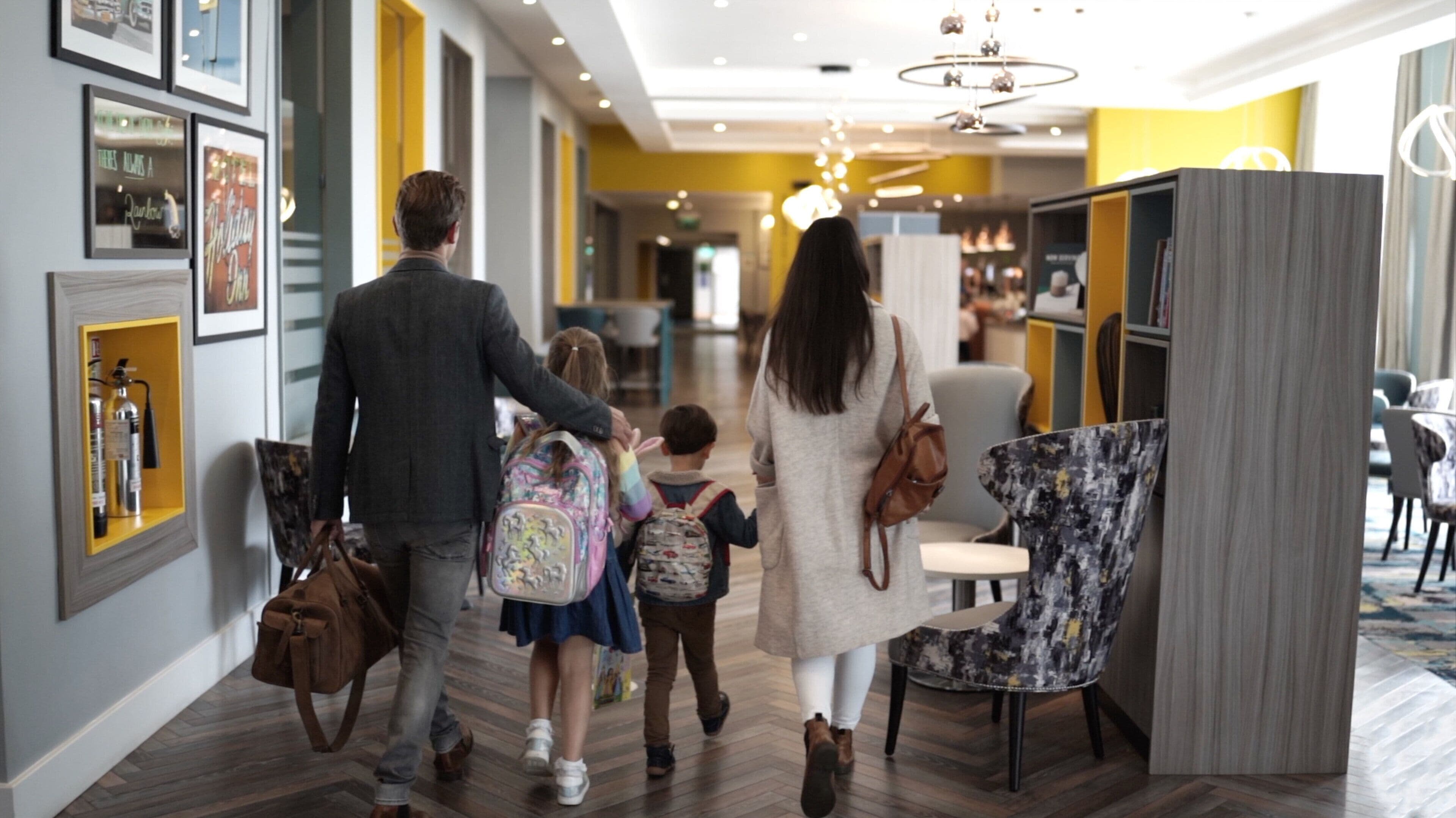 Une famille se promène dans un couloir d’hôtel, où le sol est en bois, les murs sont peints en jaune et des tableaux encadrés ornent le mur de gauche.
