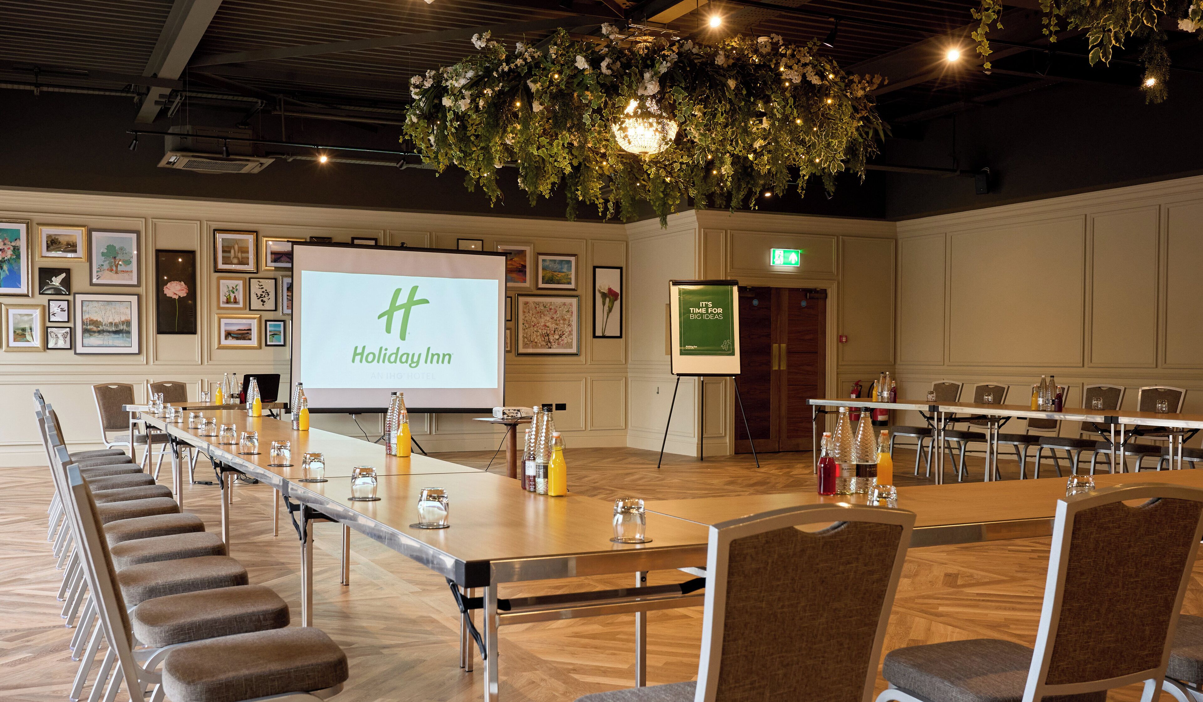 A conference room with a long table, chairs, a projector screen displaying 'Holiday Inn', and a large floral arrangement overhead.