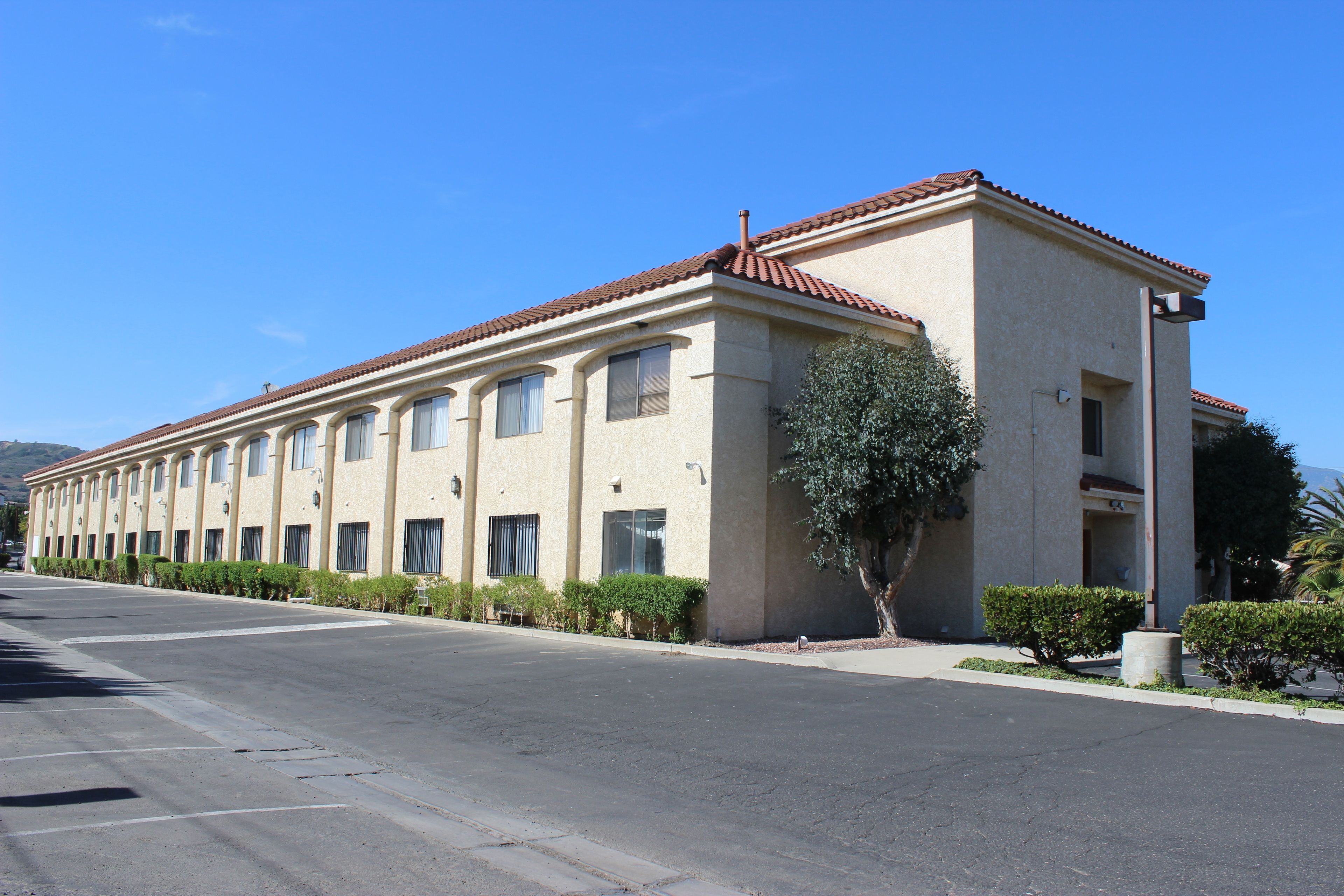 A long, beige building with a red-tiled roof, multiple windows, and a tree in front.