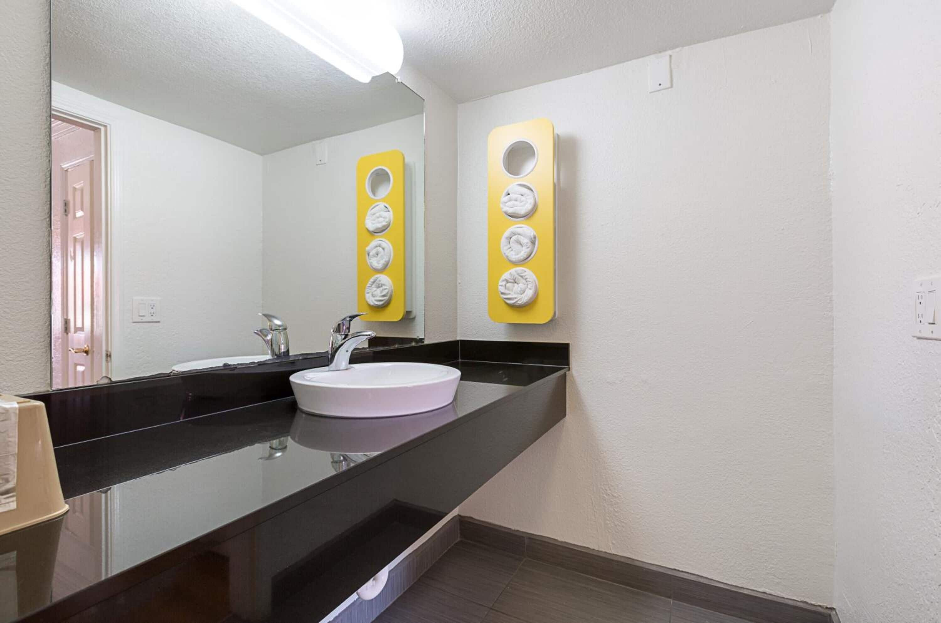 Un baño moderno con un lavamanos blanco, encimera negra y un espejo. Hay dos dispensadores de toallas amarillos en la pared.