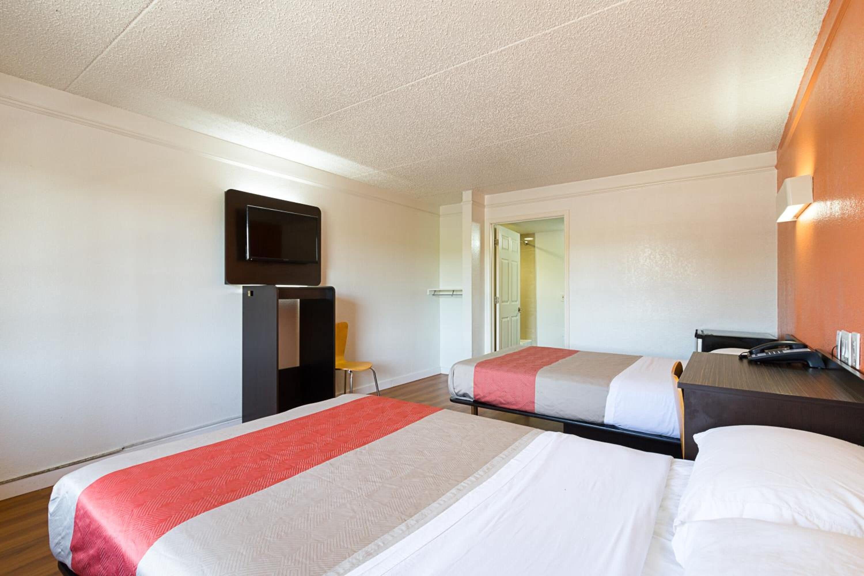A hotel room with two beds, a TV mounted on the wall, a desk with a phone, and a small table with a chair.