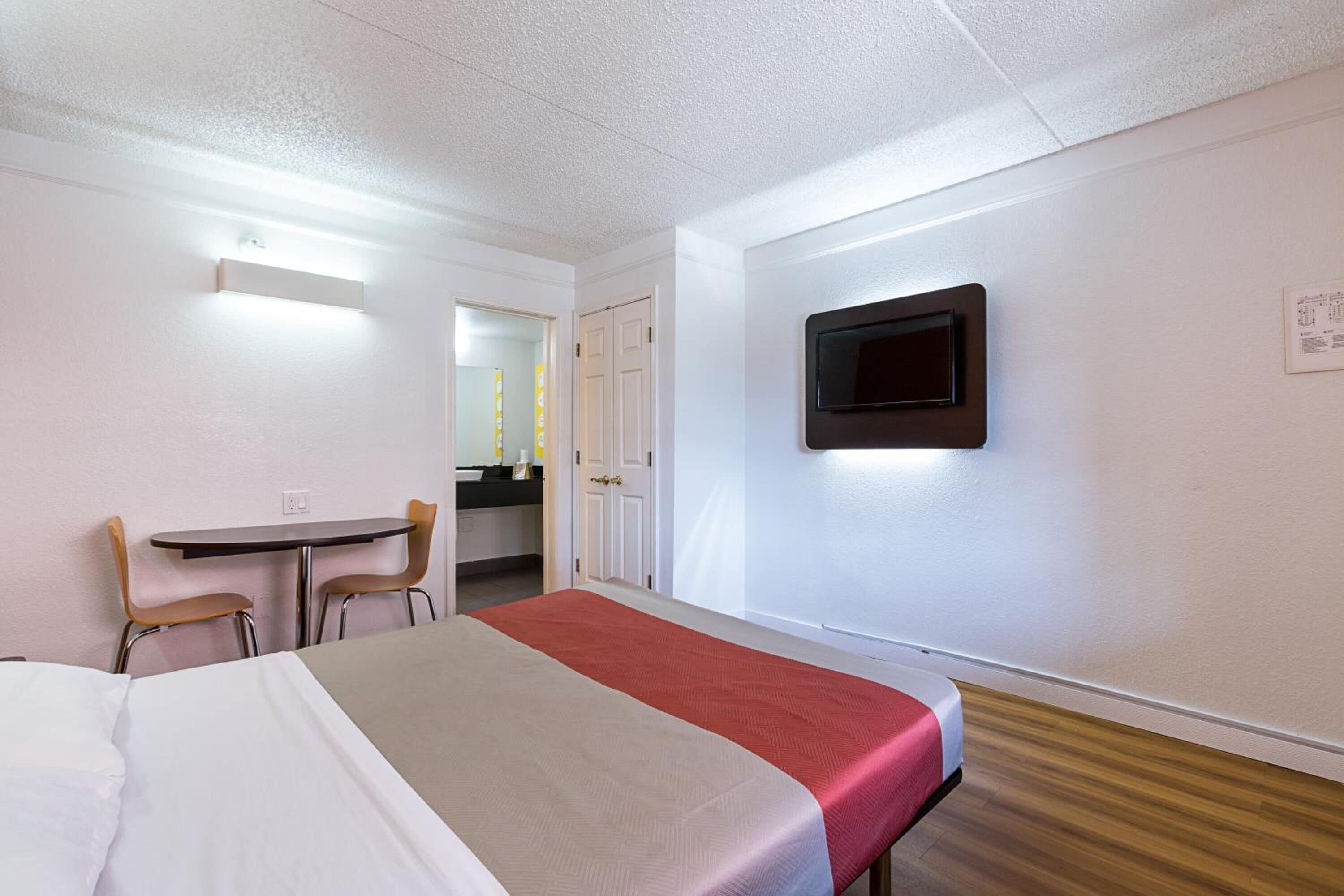 A hotel room with a bed, a desk, two chairs, a television, and a bathroom visible through an open door.