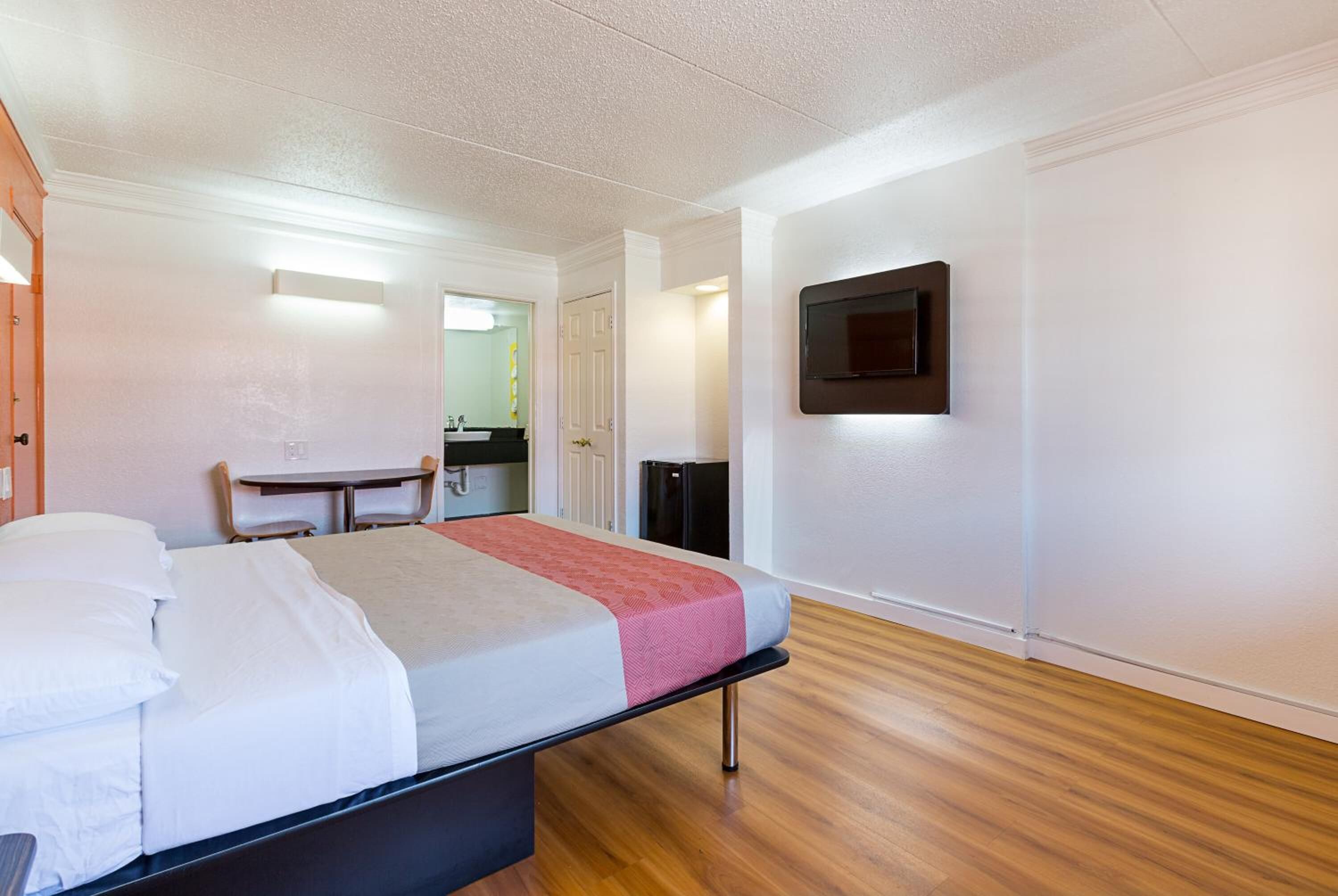 A hotel room with a bed, a desk, a chair, a TV mounted on the wall, and a door leading to a bathroom.