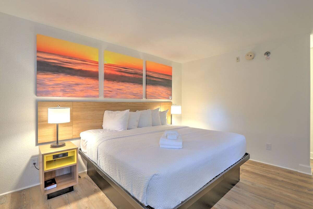 A hotel room with a large bed, two bedside lamps, a nightstand with a book, and a sunset painting above the bed.