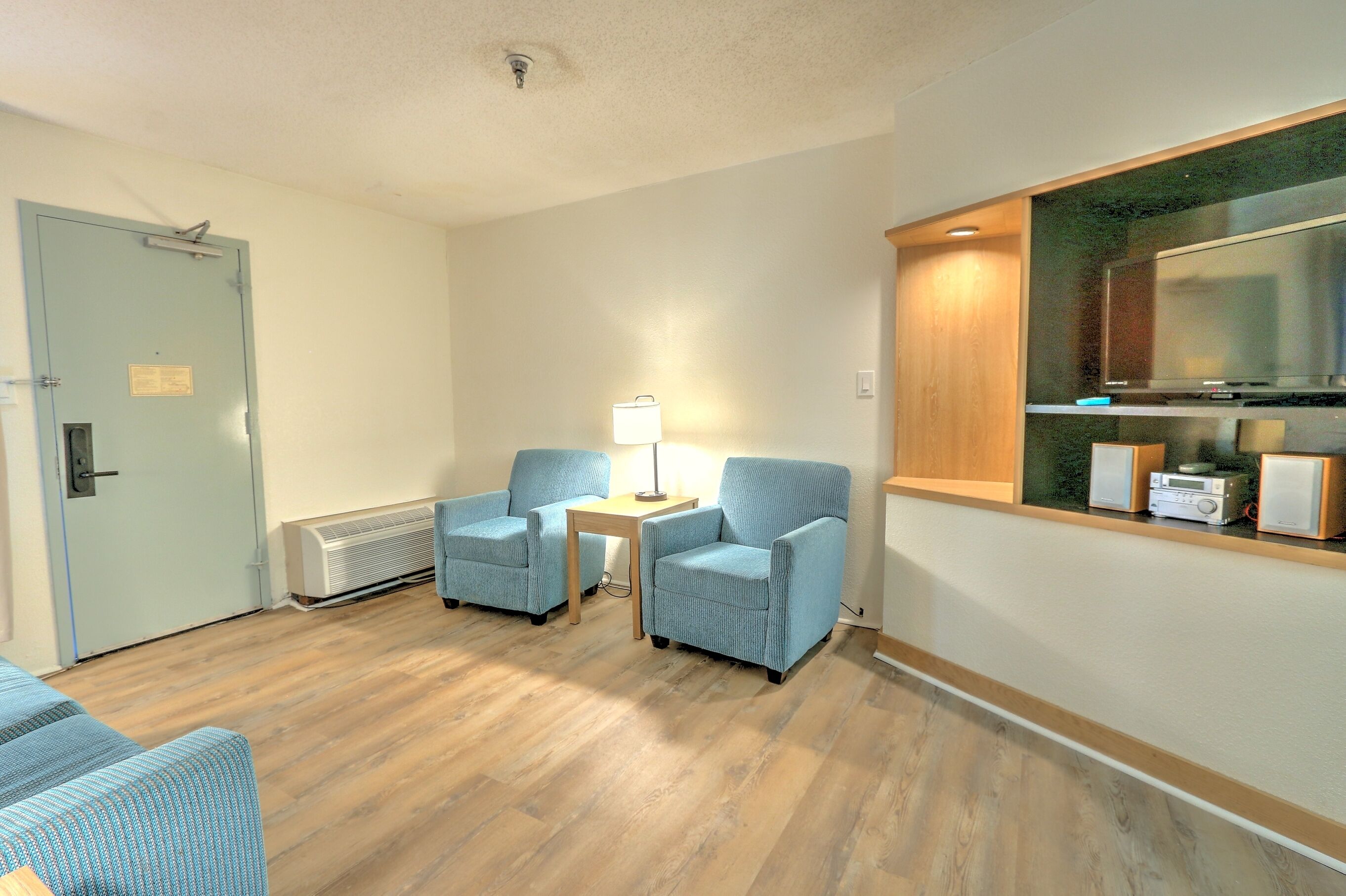 Una habitación con dos sillones azules, una mesita con lámpara, una puerta de madera y una kitchenette con microondas y tostadora.
