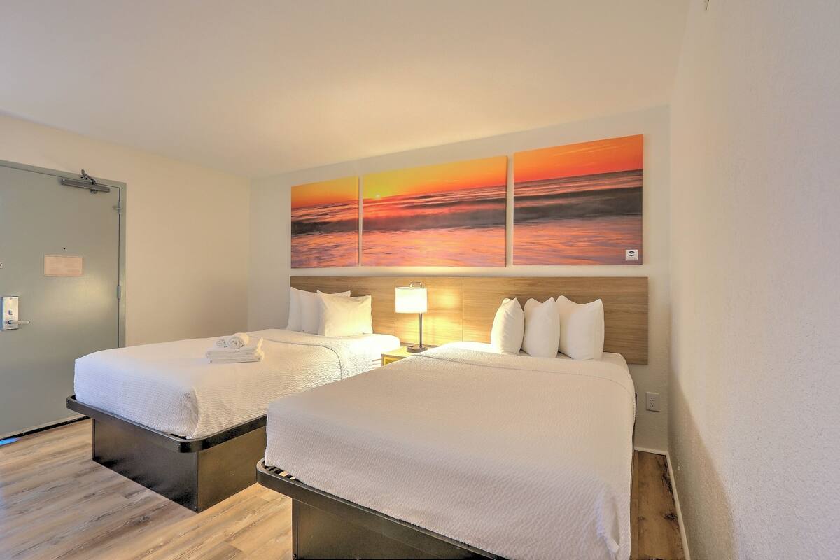 A hotel room with two beds, a wooden headboard, a painting of a sunset, and a nightstand with a lamp.
