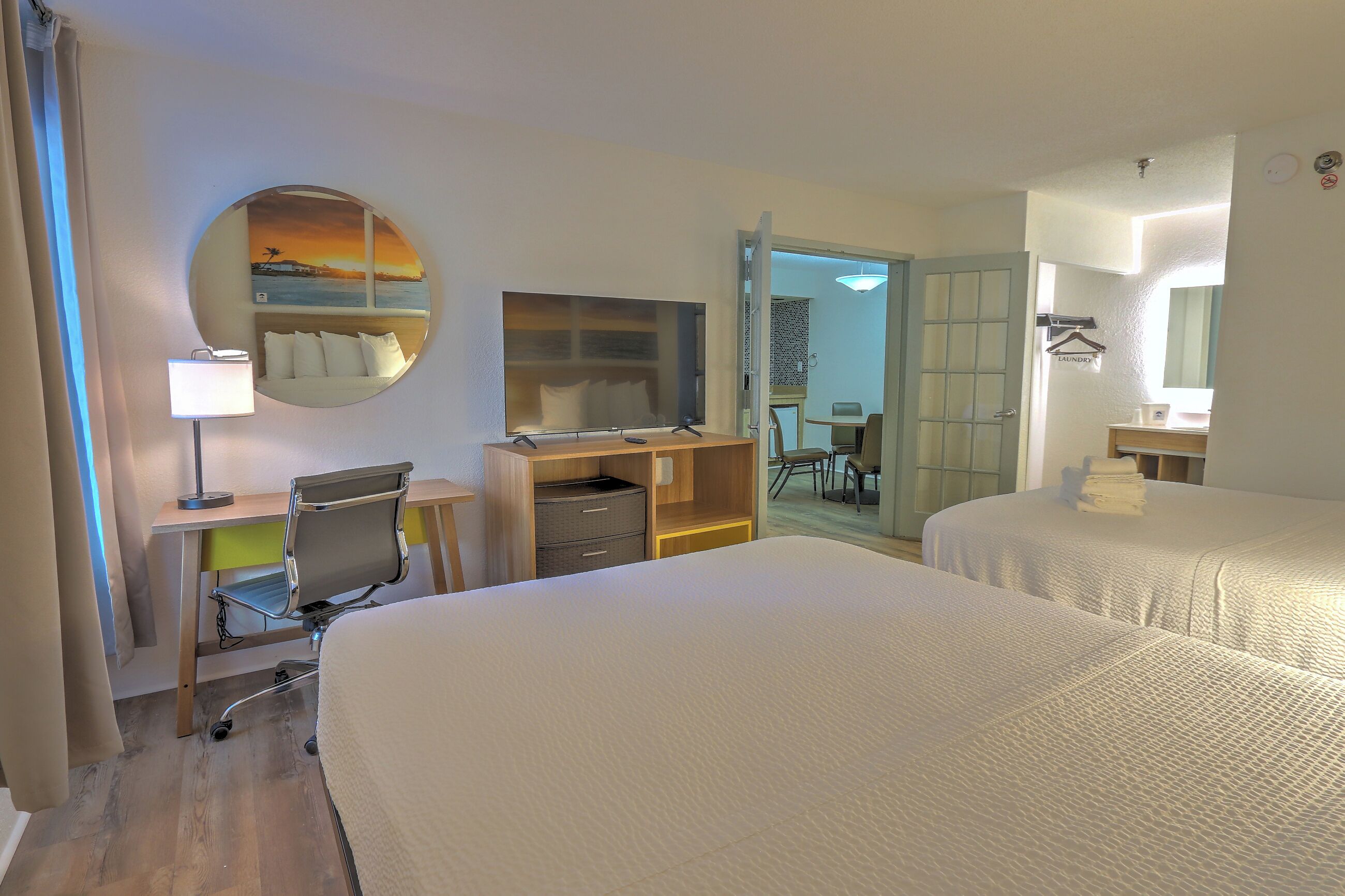 A hotel room with two beds, a desk with a chair, a round mirror, a TV, and a view of the outside.
