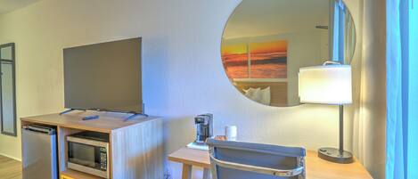 Een kleine hotelkamer met een kitchenette, een flatscreen televisie, een bureau met lamp, een stoel en een grote spiegel die uitzicht op een ondergaande zon weerspiegelt.